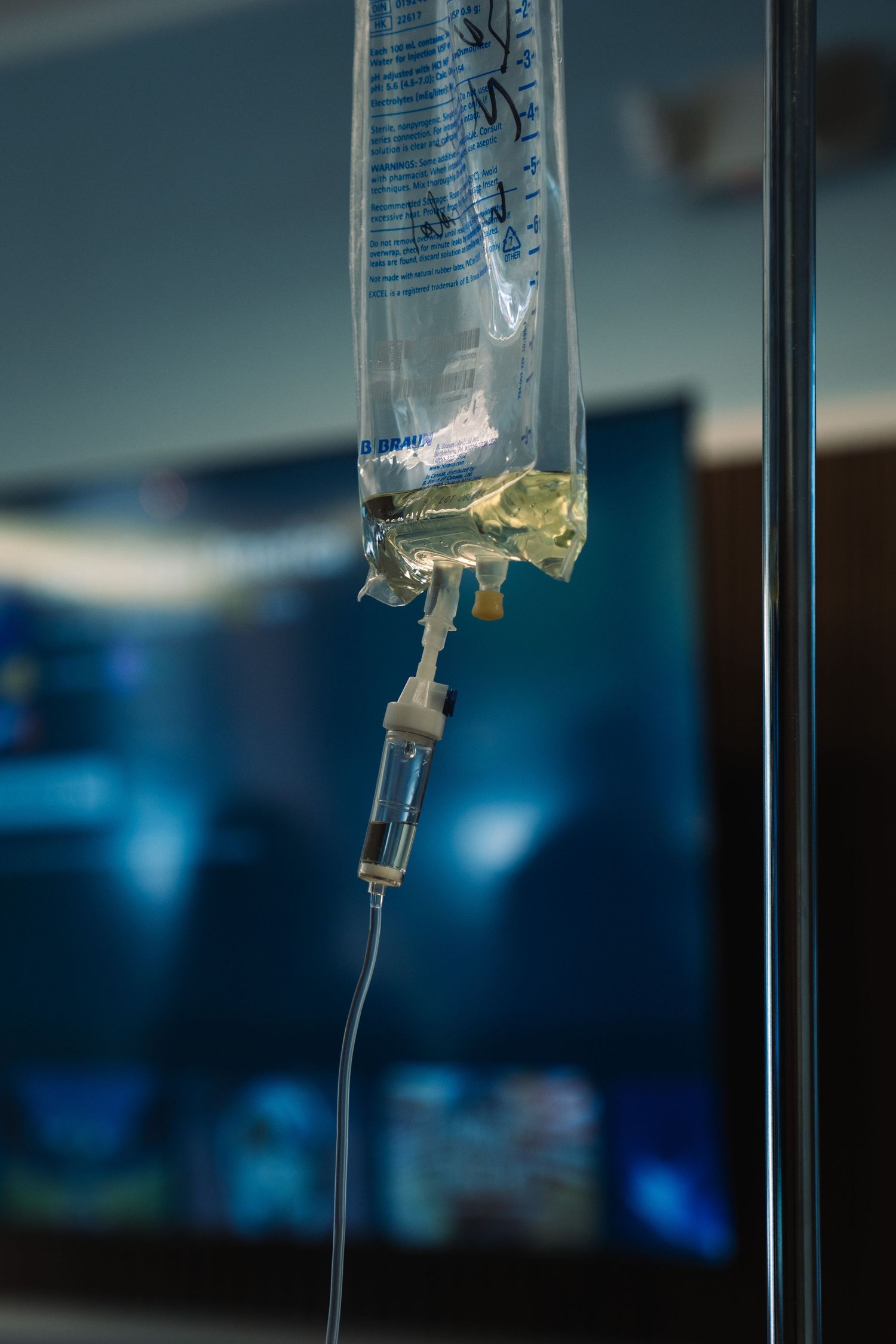 Close-up of a medical IV drip bag with tubing hanging on a pole in a hospital room with a blurred TV screen background.