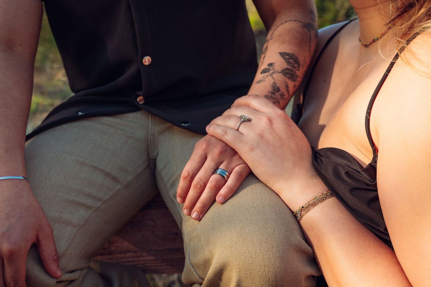 Close-up of a couple holding hands, showing a blue ring and bracelet, with a tattooed arm resting gently on top.