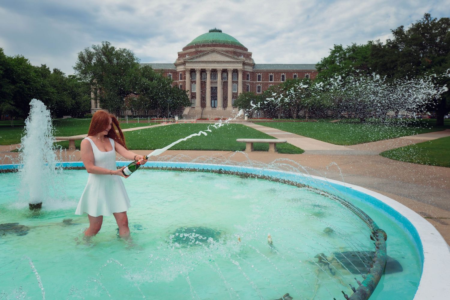 Woman in white dress standing in a fountain, spraying water near a domed university building on a sunny day.