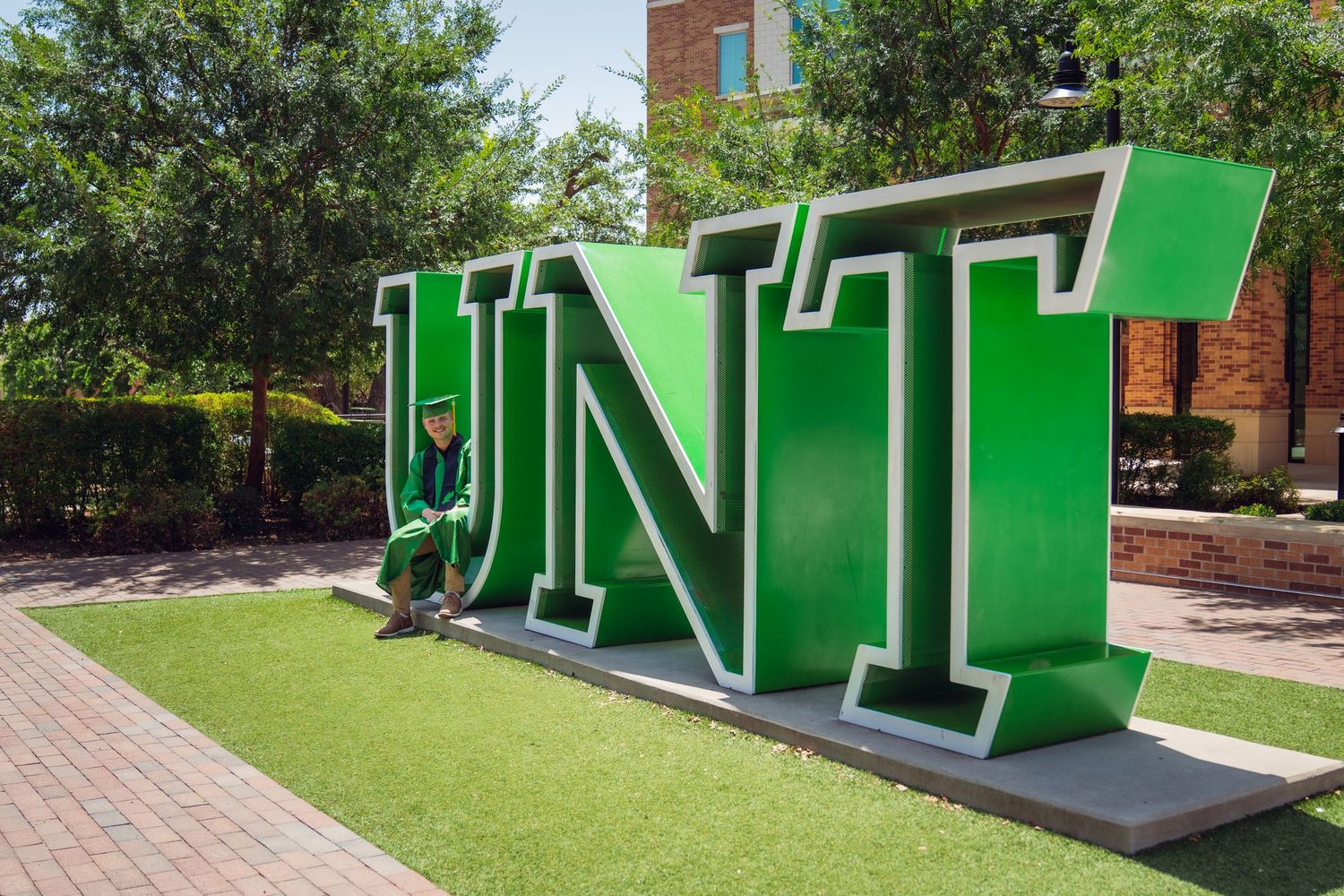 Graduate in cap and gown posing beside large green UNT letters sign on University of North Texas campus.