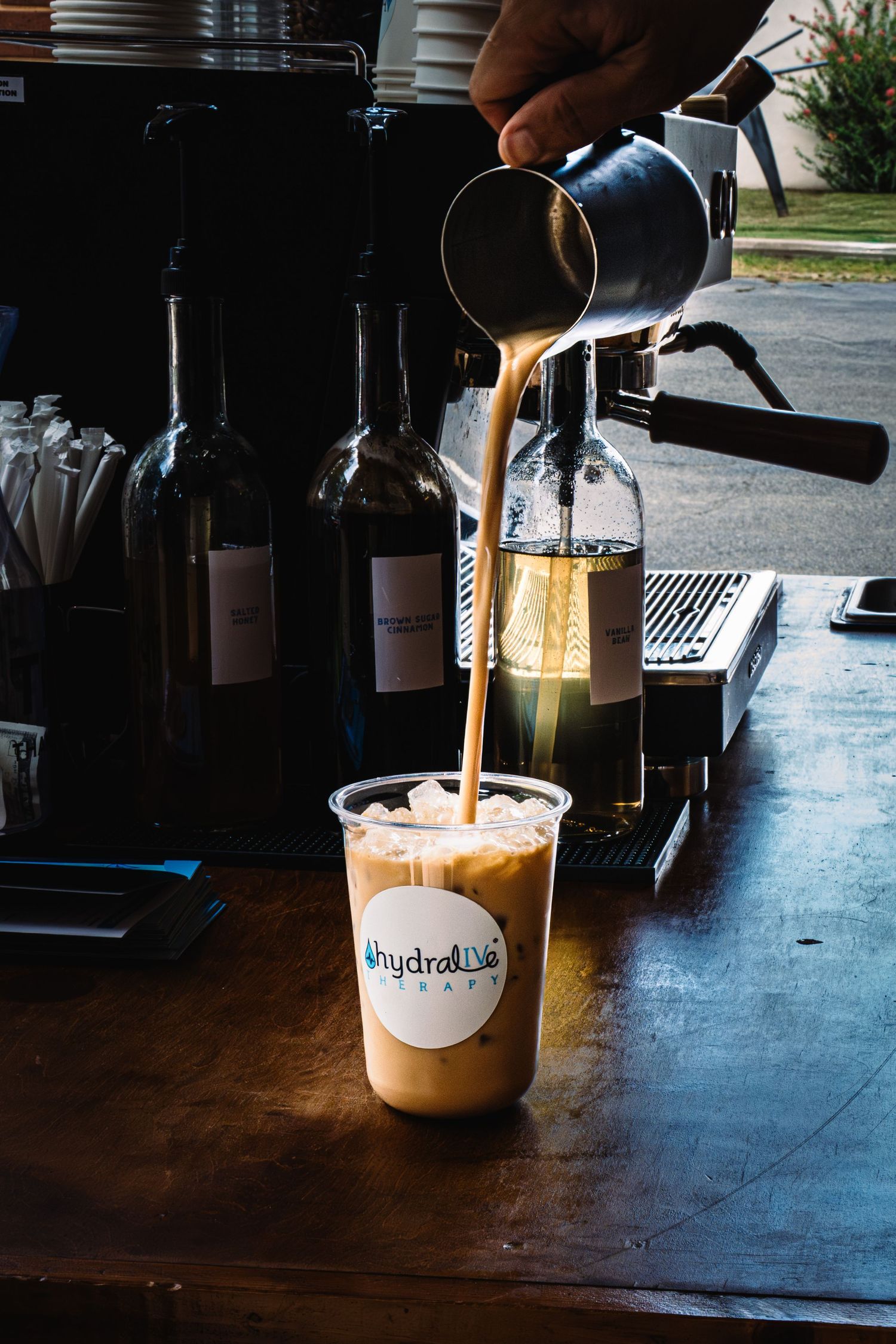 Barista pouring espresso into an iced coffee cup at Hydralise Cafe with bottles and espresso machine in background.