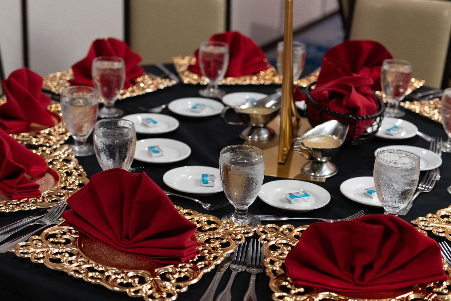 Elegant round banquet table with black tablecloth, gold lace overlay, red folded napkins, and crystal glassware for formal dining.