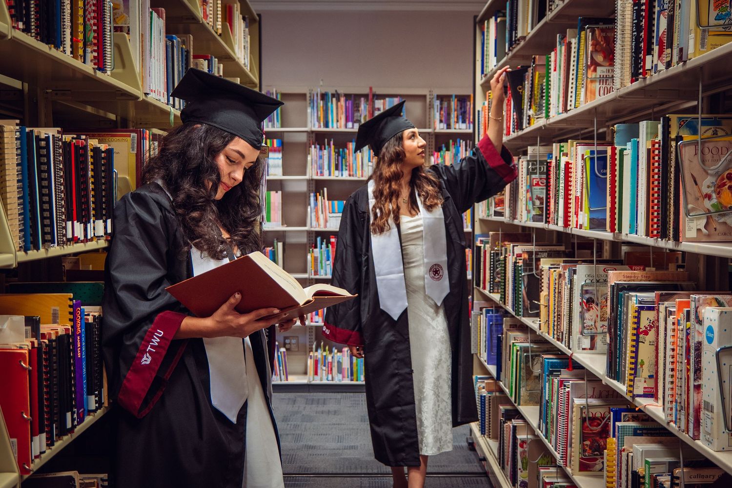 Two female graduates in black caps and gowns browsing books in a university library.