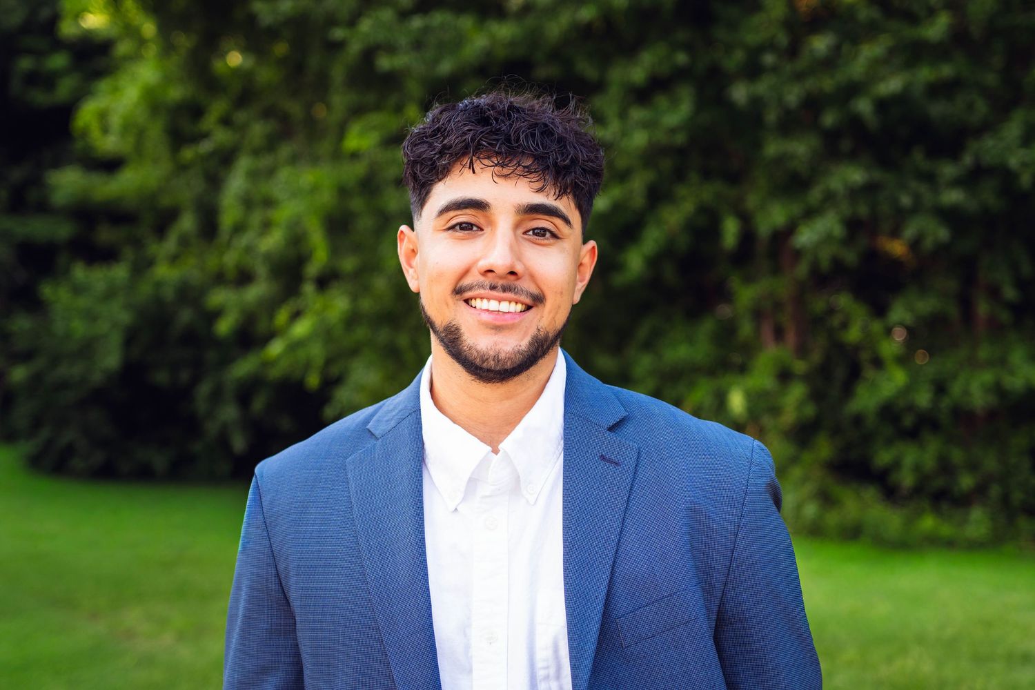 Young man in blue blazer smiling outdoors with green foliage in the background.