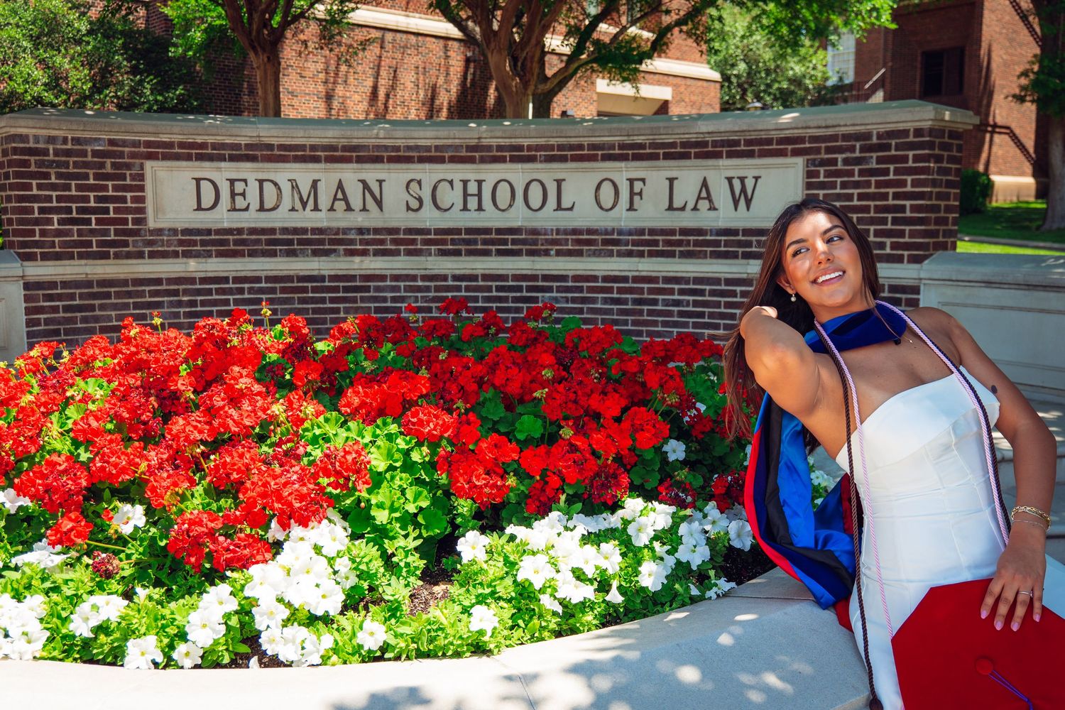 Smiling graduate in blue regalia posing by Dedman School of Law brick sign.