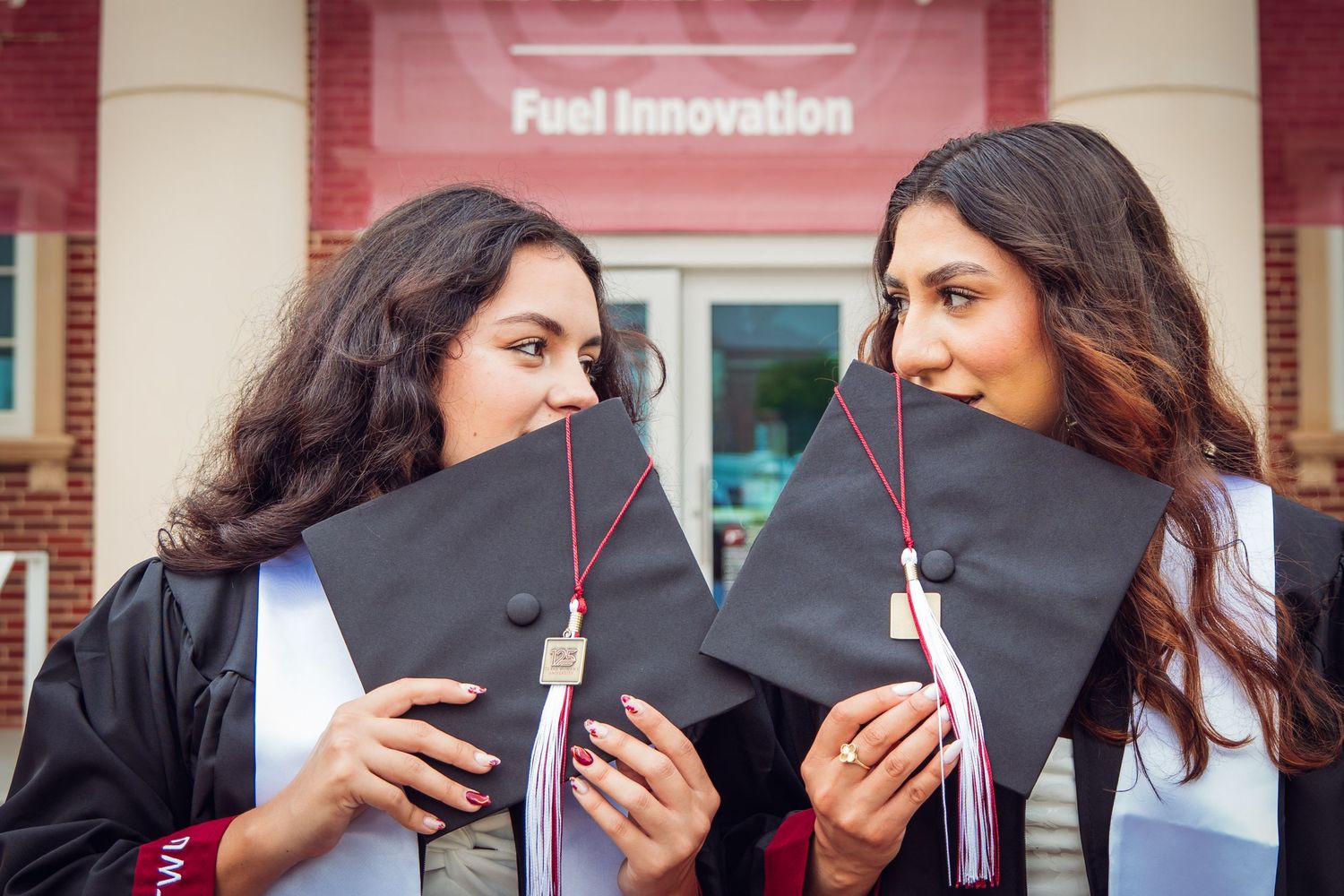 Two graduates hiding faces behind black mortarboards in front of Fuel Innovation sign.