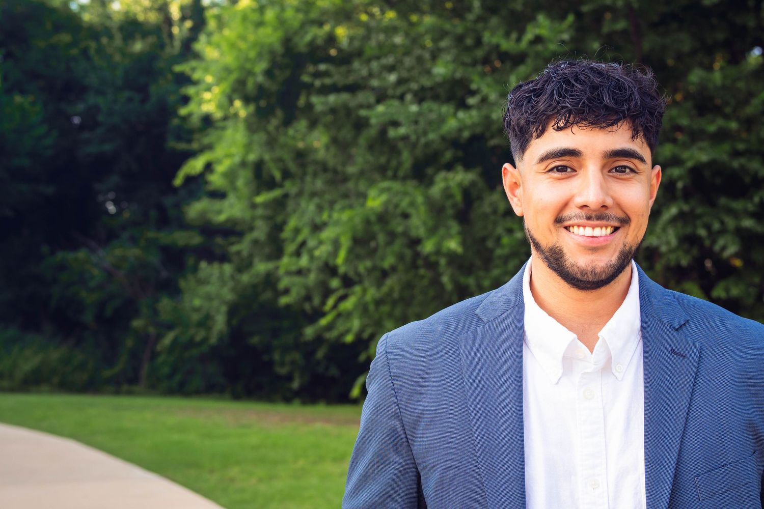 Smiling young man in a blue blazer and white shirt posing outdoors with green trees in the background.