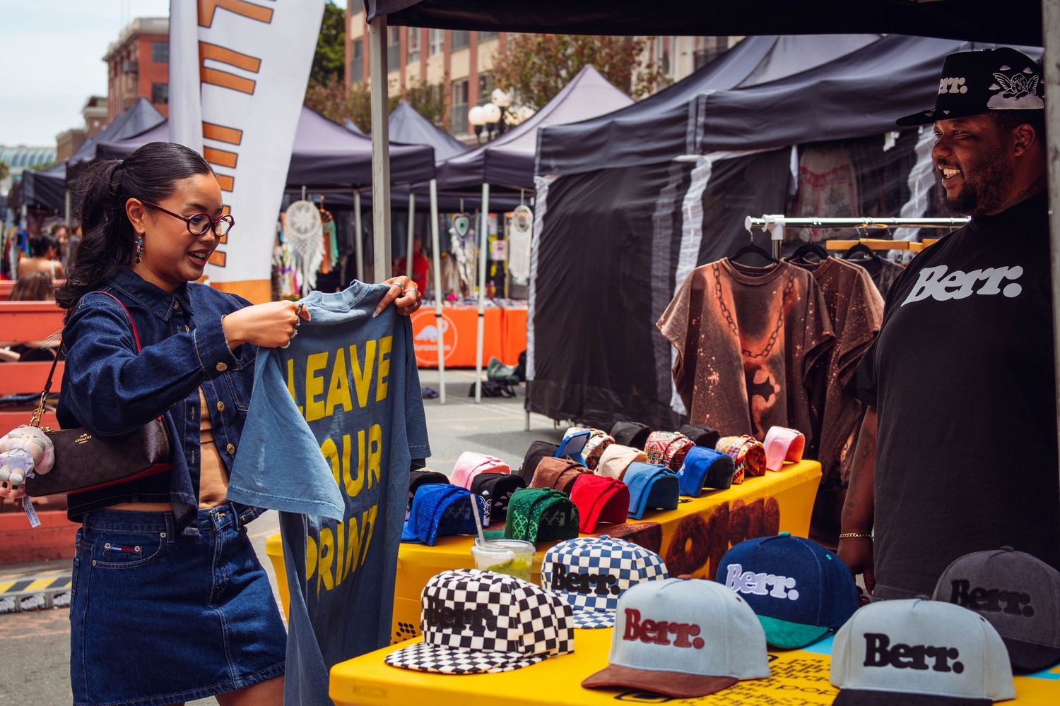 Young woman browsing hats and apparel at an outdoor street market vendor booth.