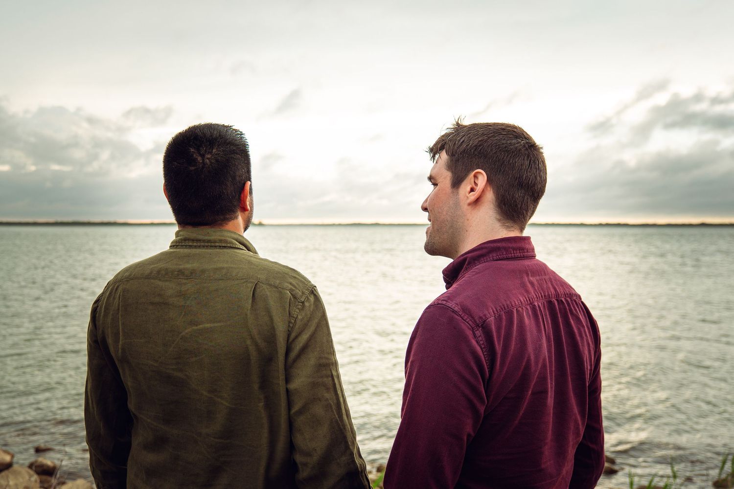 Two men standing with their backs turned, gazing at a calm lake under a cloudy sky.