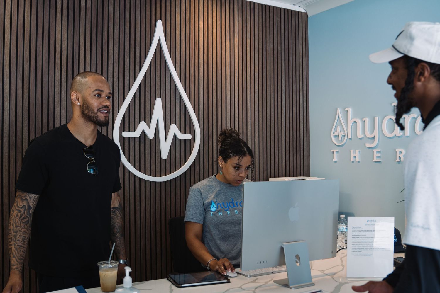 Three people chat at a modern hydration therapy clinic reception desk with a water drop logo on the wall.