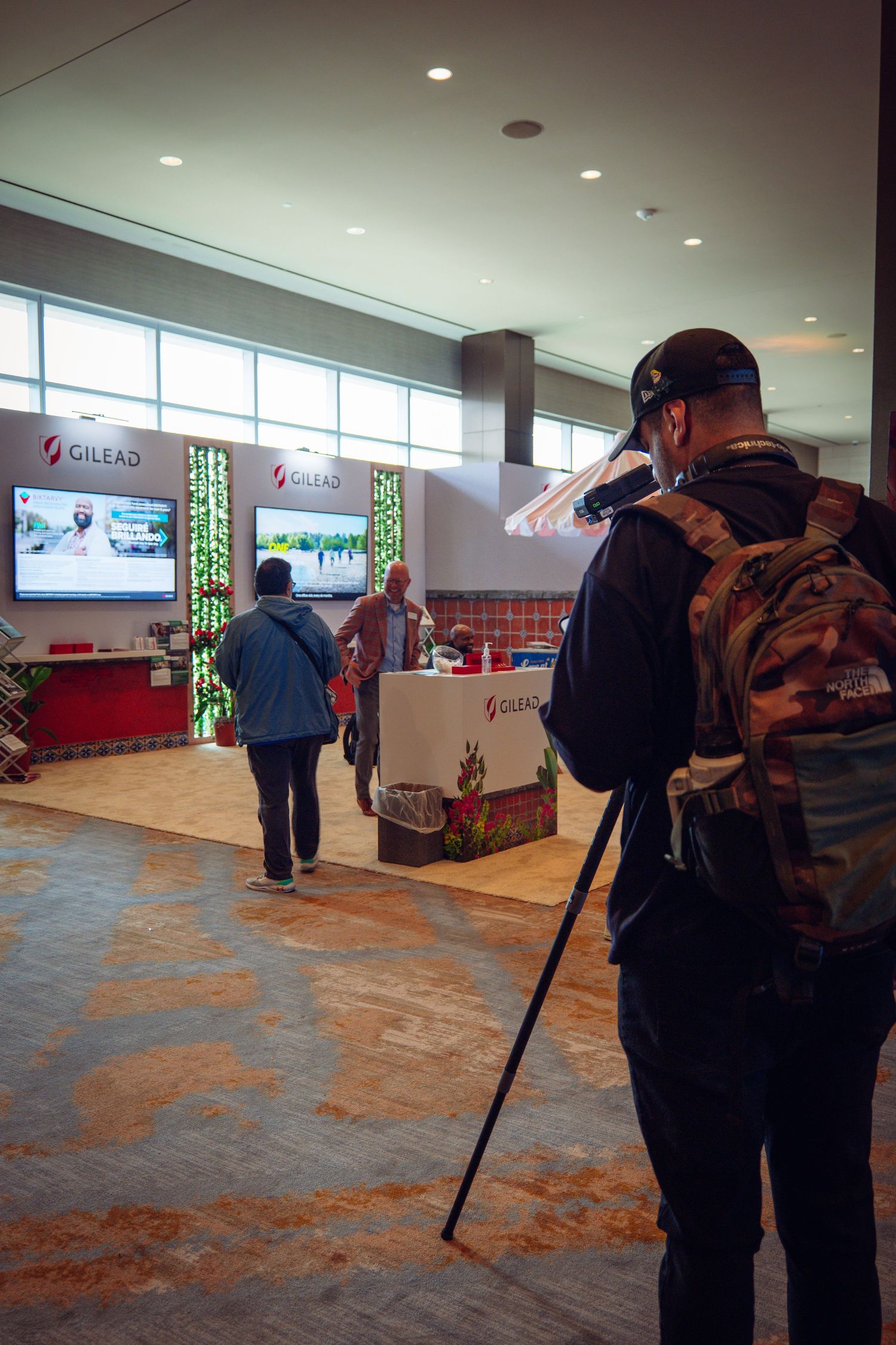 Photographer with tripod captures visitors at a Gilead Sciences exhibition booth inside a convention center.