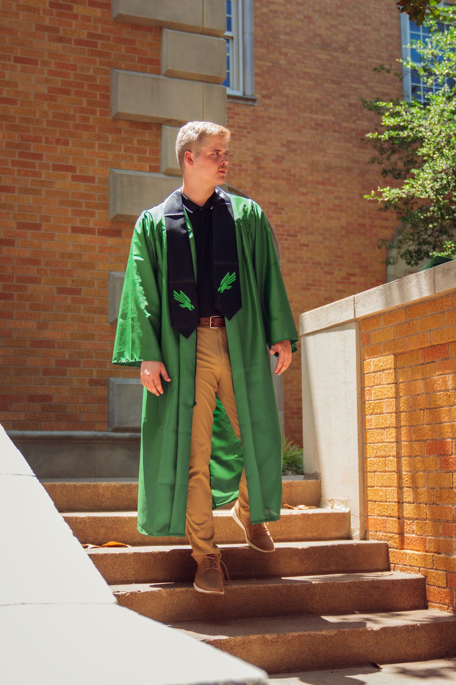 Man in green graduation gown standing on outdoor steps near brick building on a sunny day.