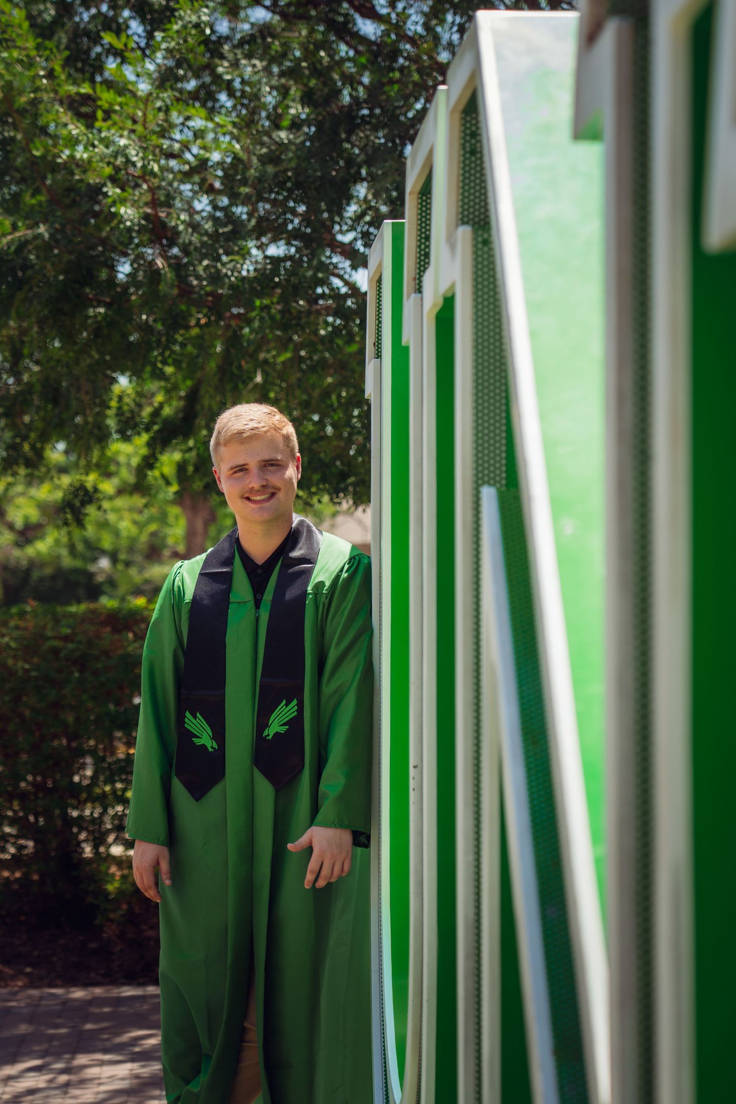 Young man smiling in green graduation gown standing outdoors near a green and white striped wall.