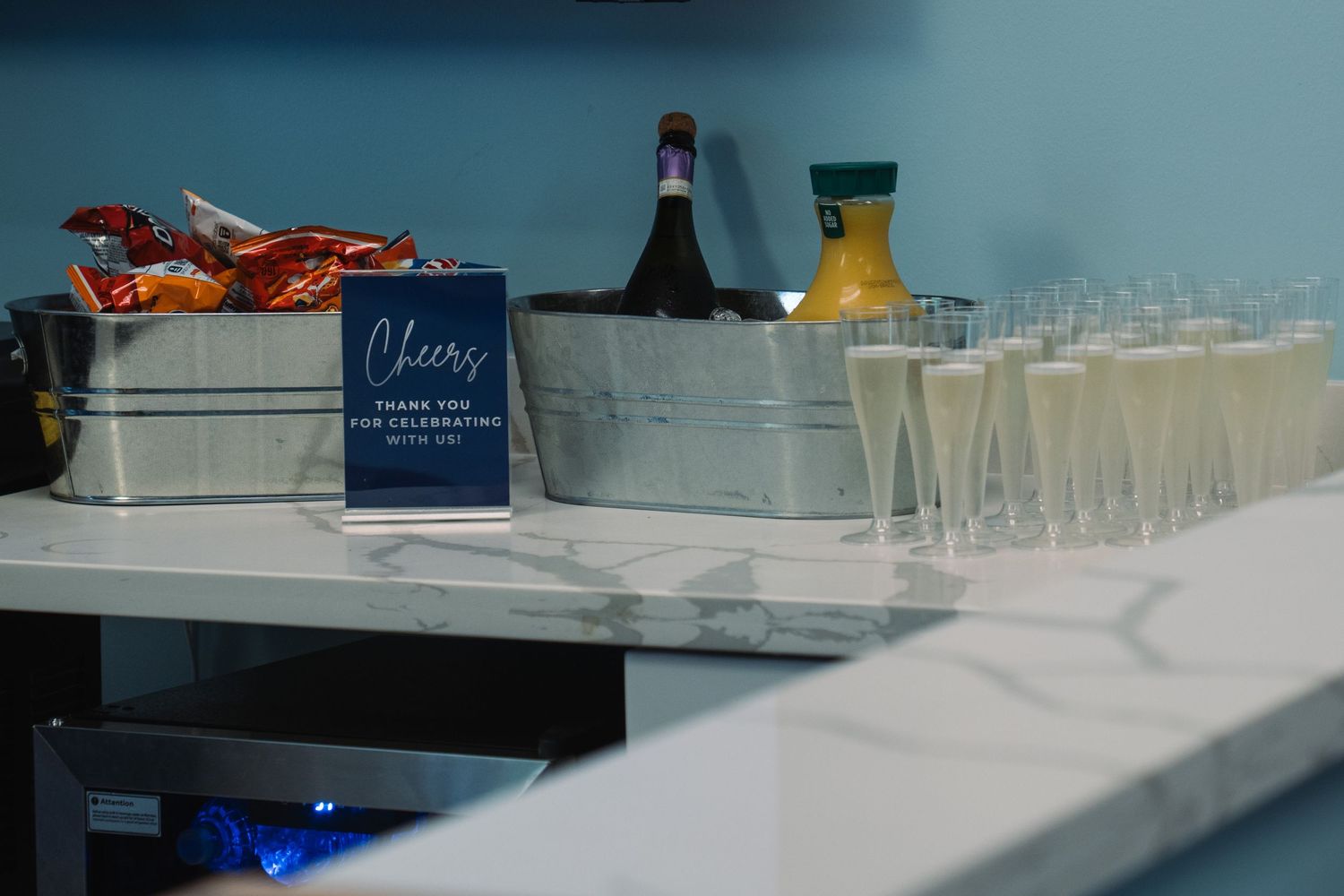 Elegant champagne flute display with Cheers sign, orange juice, and snacks on marble bar for celebration event.