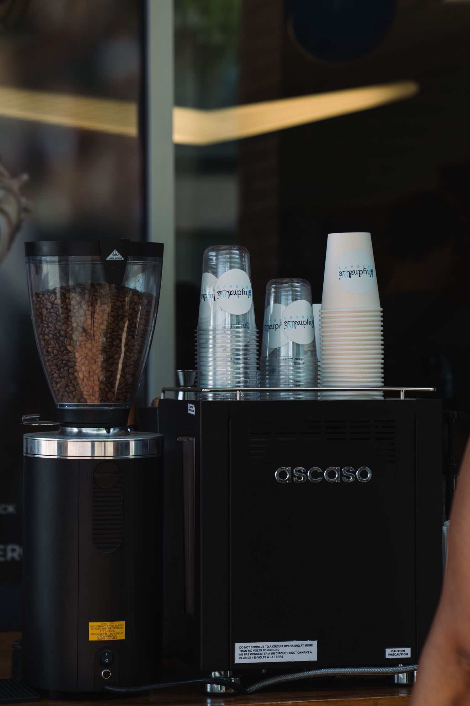 Ascaso espresso machine with coffee bean grinder and stacked paper cups on a cafe counter.