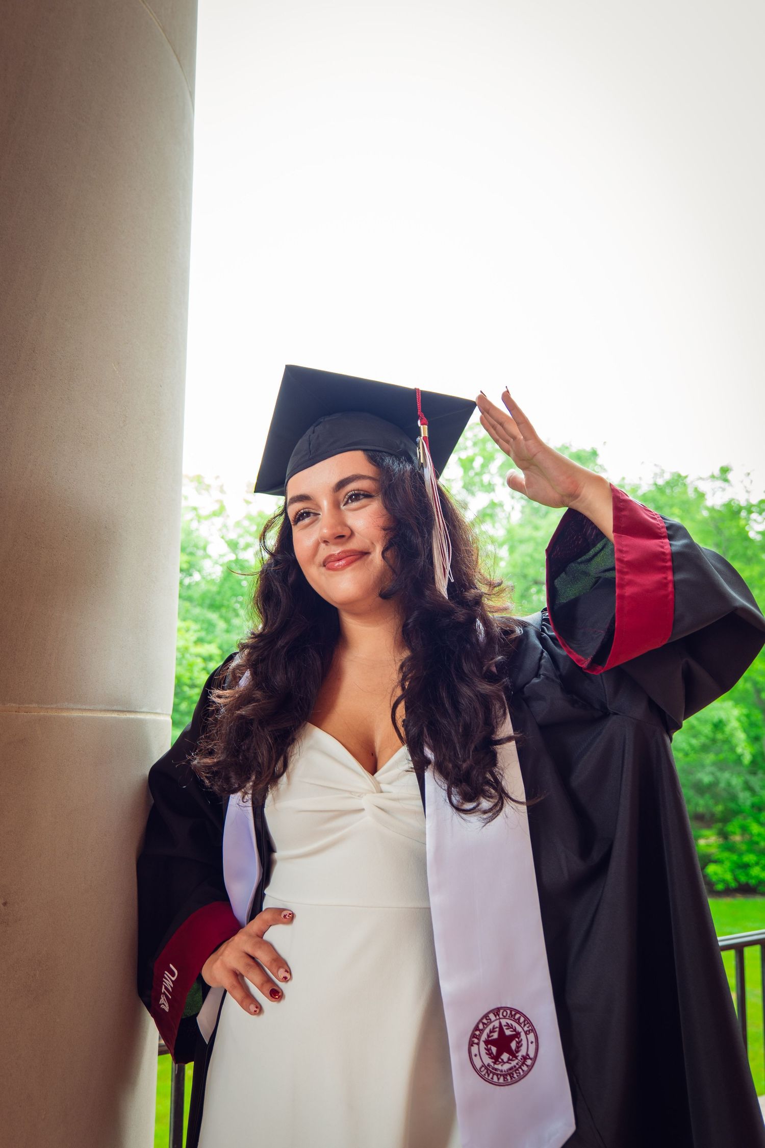 Smiling graduate in black cap and gown with white dress leaning against column outdoors.