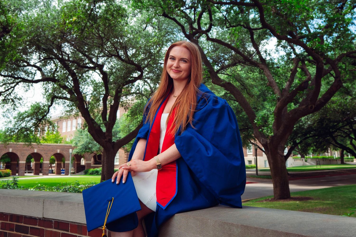 Young woman in blue graduation gown and cap sitting outdoors on campus surrounded by trees on a sunny day.