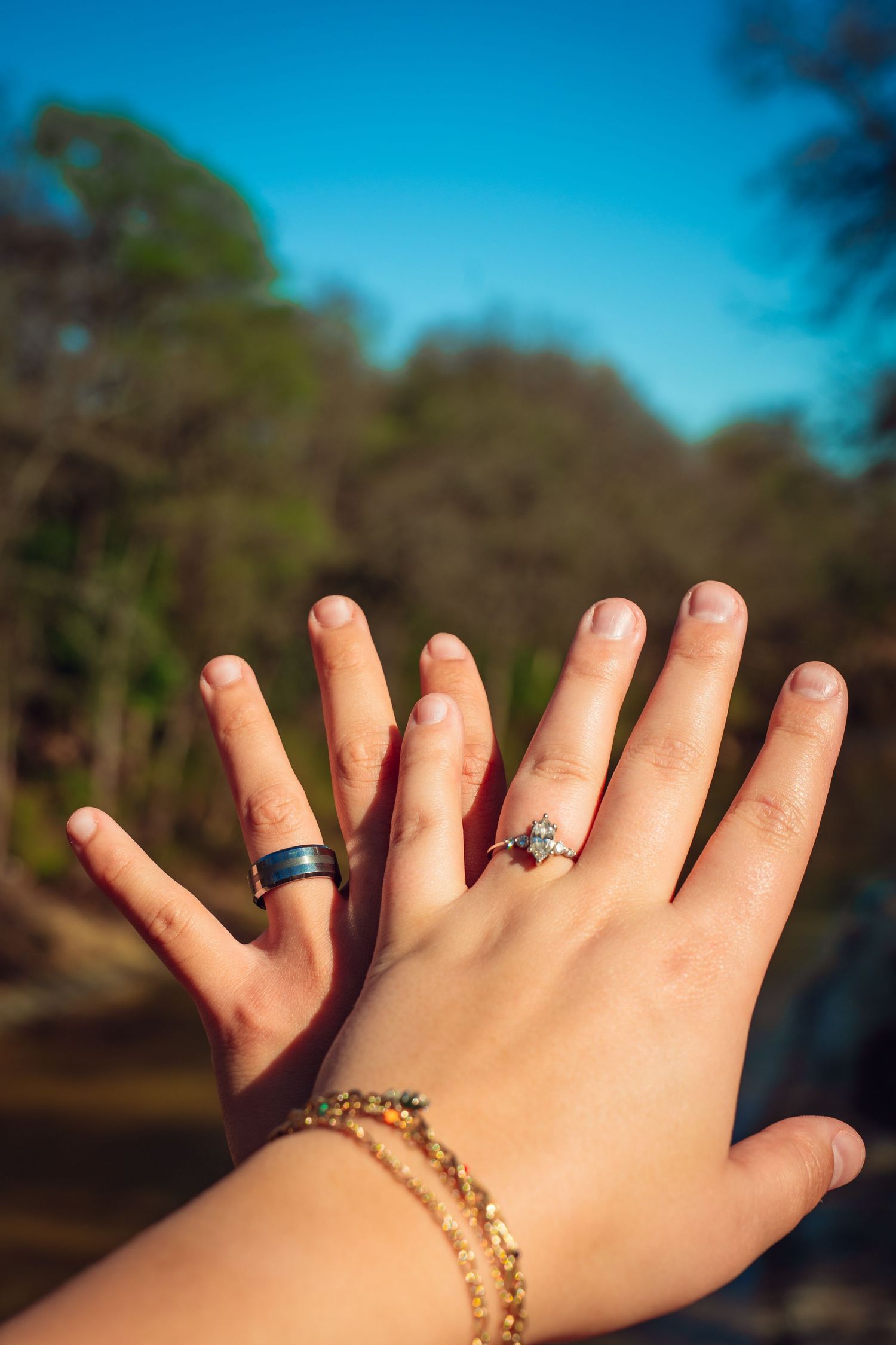 Two hands raised outdoors showing matching engagement rings, one with a diamond ring and bracelet, against a blue sky backdrop.