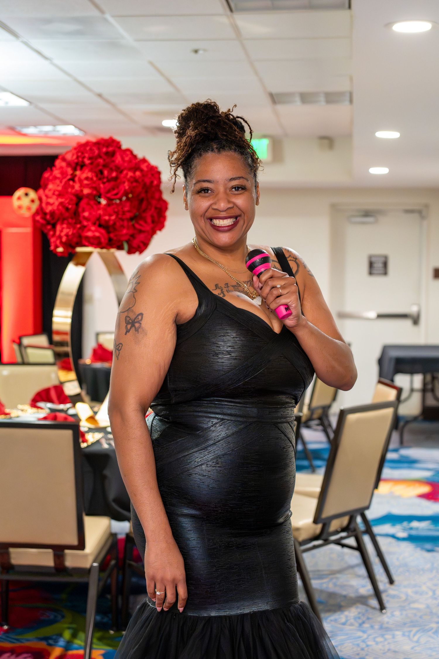 Woman in black leather dress holds pink microphone at elegant event with red floral centerpieces.