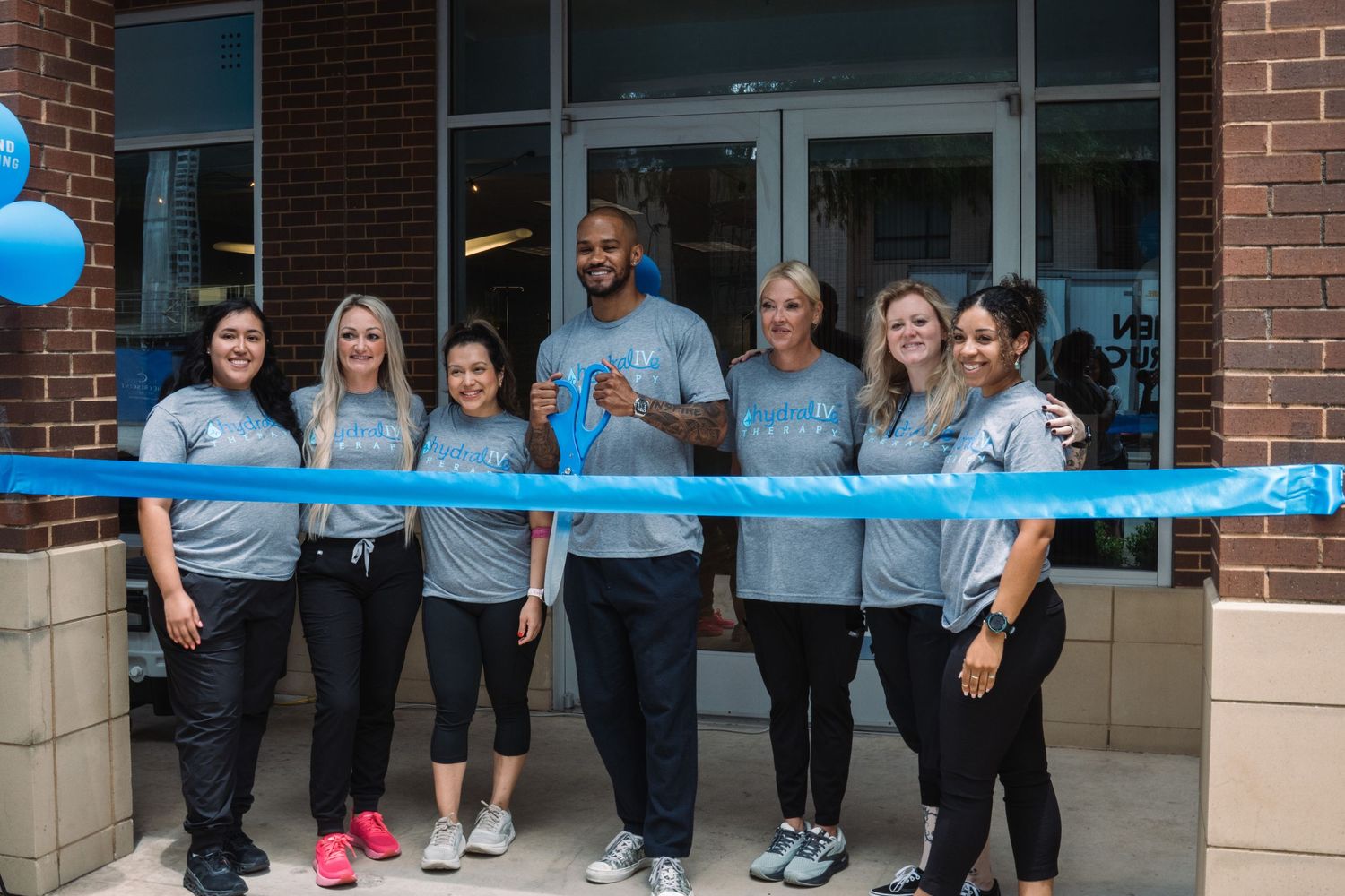 Group of seven smiling staff members in gray t-shirts cutting a blue ribbon at a grand opening ceremony outside a brick building.