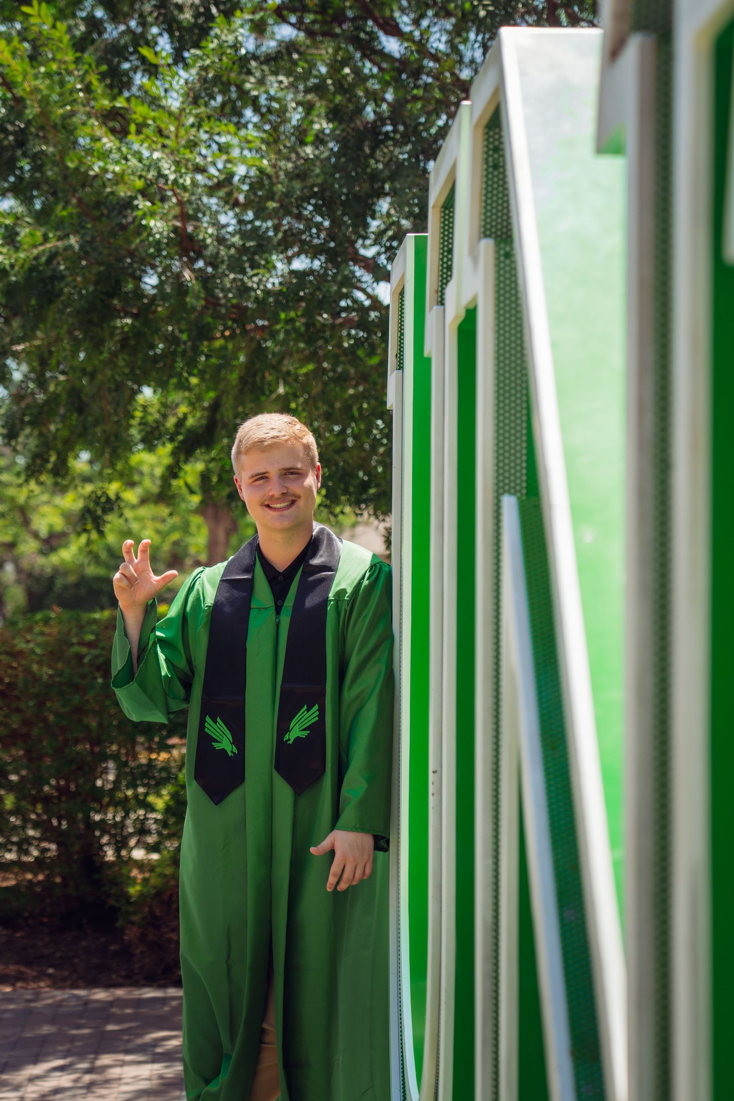 Young man in green graduation gown and stole waving beside a green and white structure outdoors.