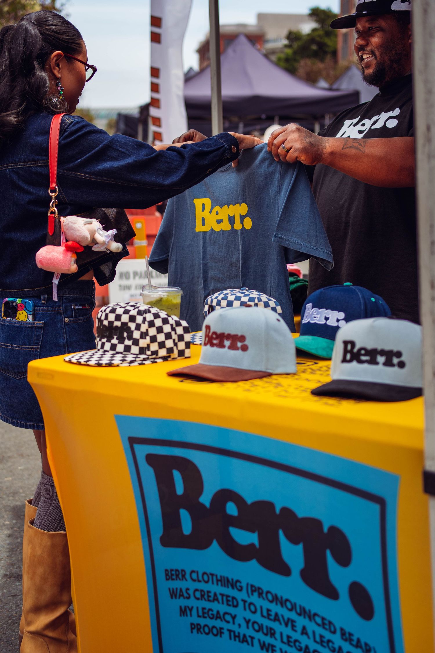 A vendor displays Berr. clothing brand merchandise including t-shirts and snapback hats at an outdoor market stall.