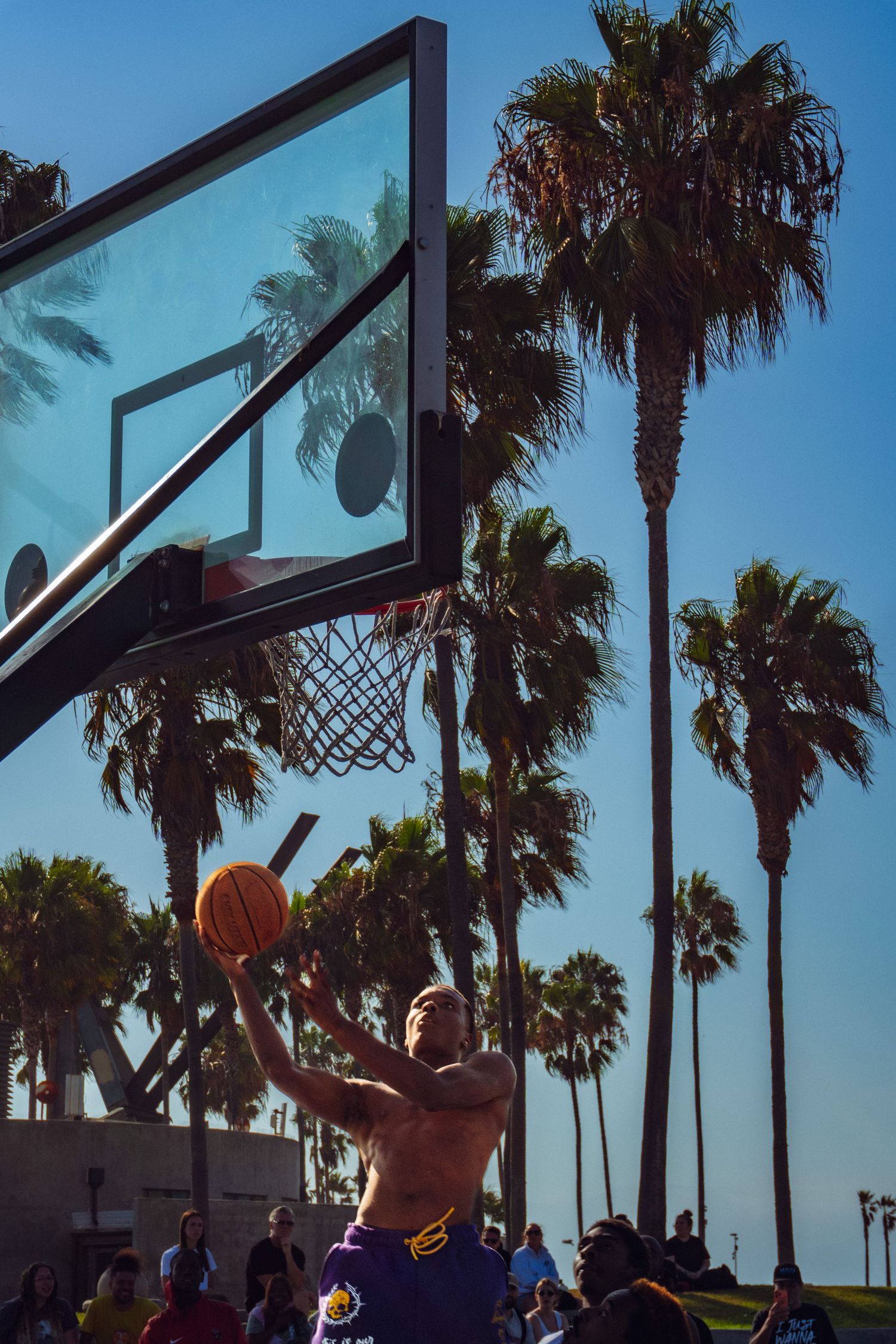 Shirtless man shooting basketball at outdoor court with palm trees and blue sky at Venice Beach.