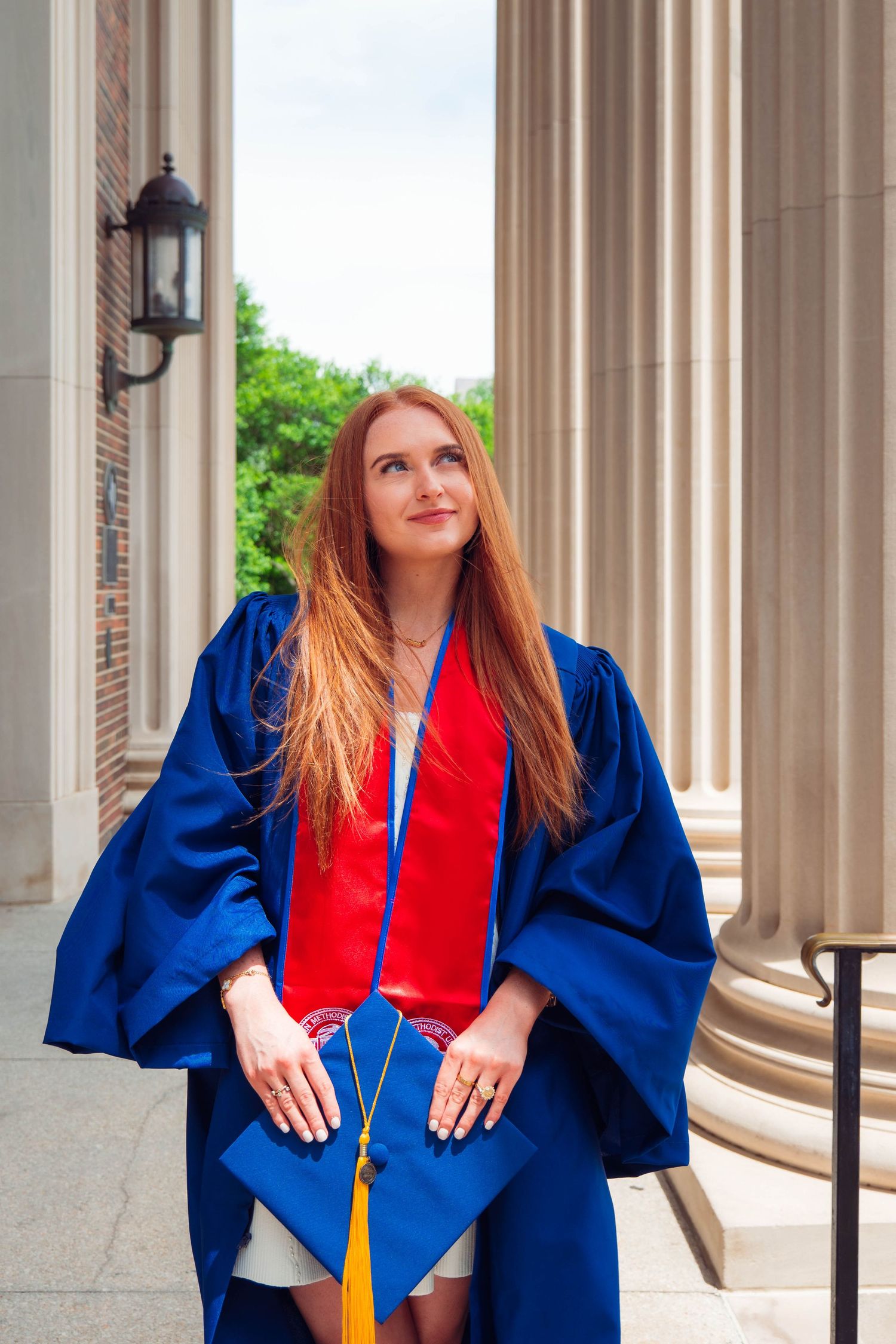Young redhead woman in blue graduation gown holding cap while looking up between columns.