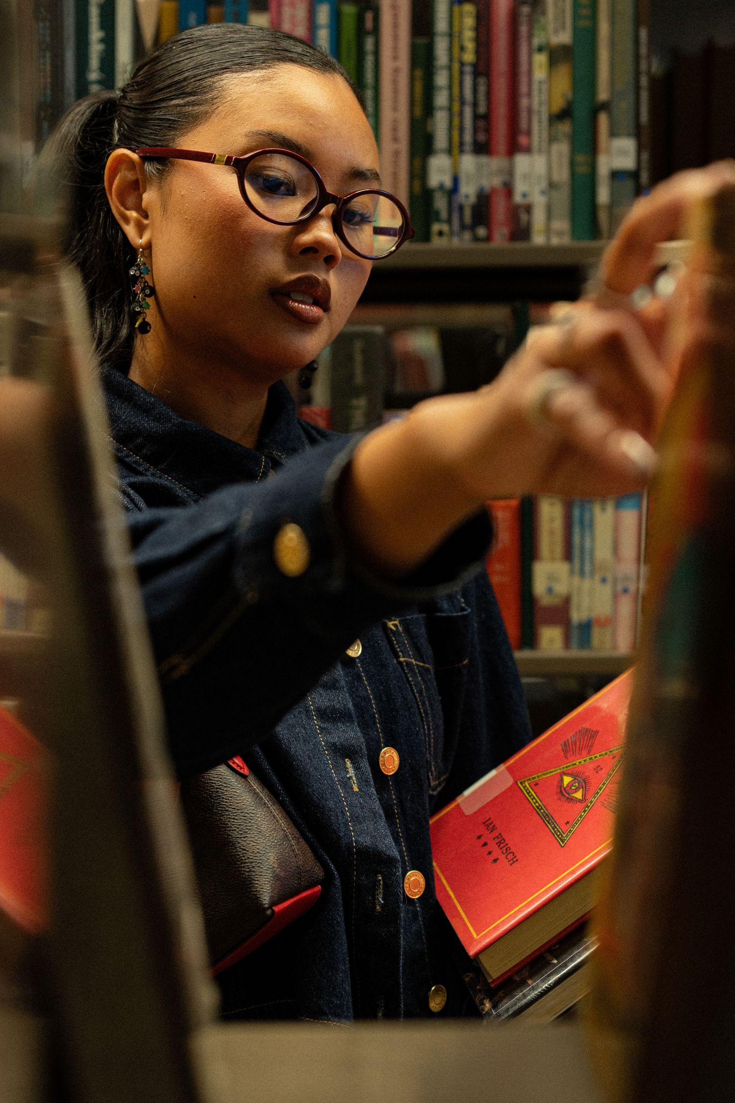 Young woman with glasses browsing books in a library, reaching for a title on a shelf.