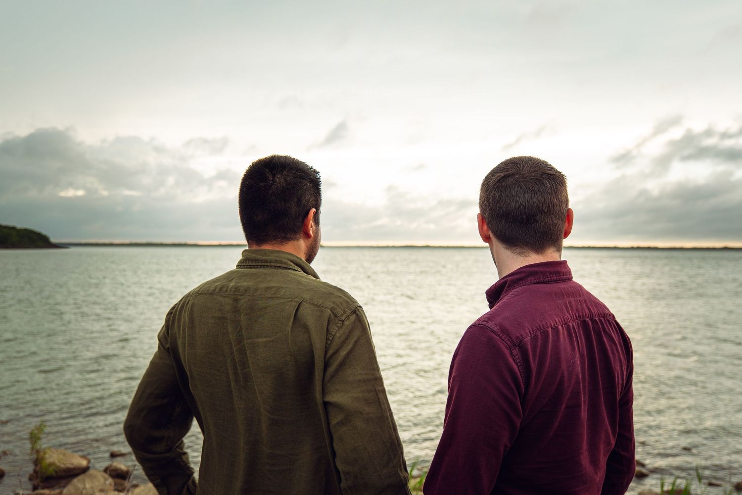 Two men standing side by side looking out at a calm lake under a cloudy sky.