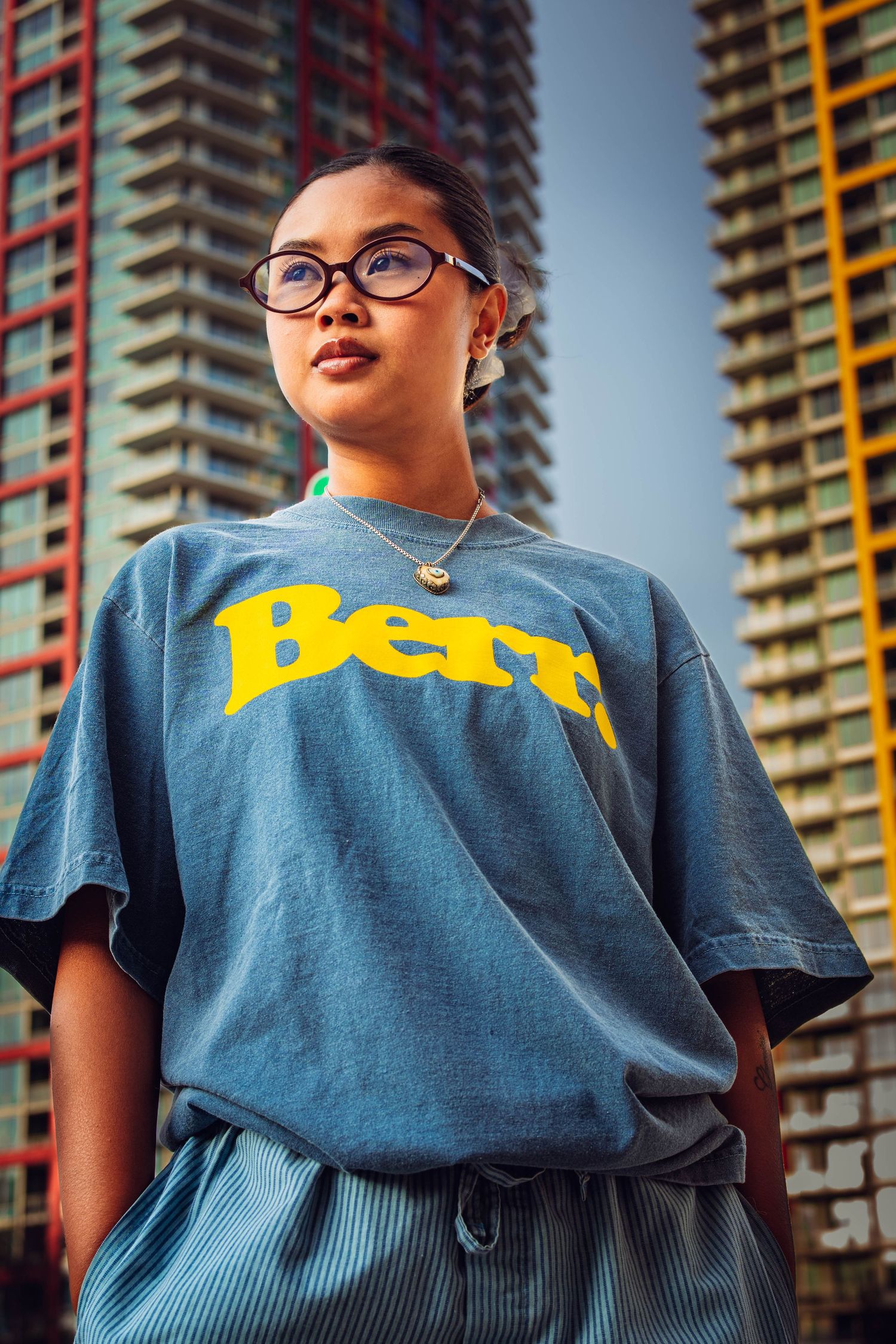 Young woman in blue Berr. graphic tee and glasses poses confidently in front of urban high-rise buildings.