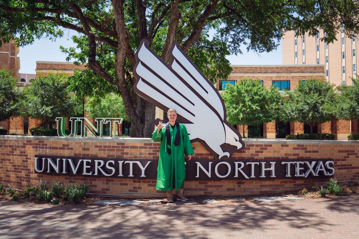 Graduate in green cap and gown poses proudly in front of University of North Texas sign with eagle sculpture.