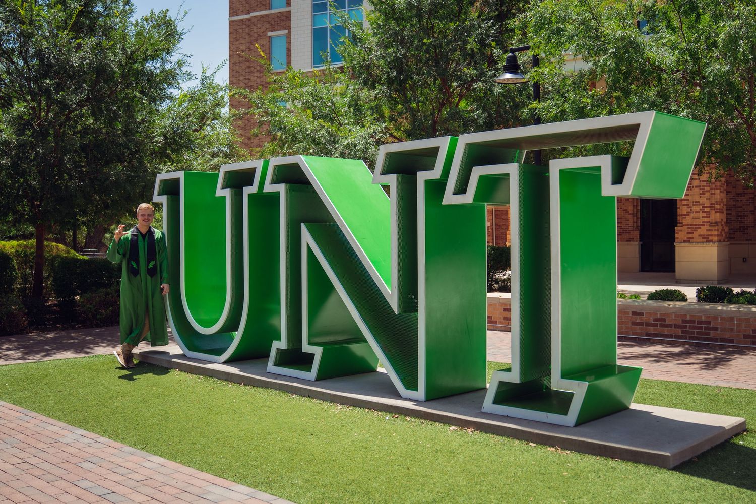 Graduate poses beside large green UNT block letters sculpture on University of North Texas campus on a sunny day.