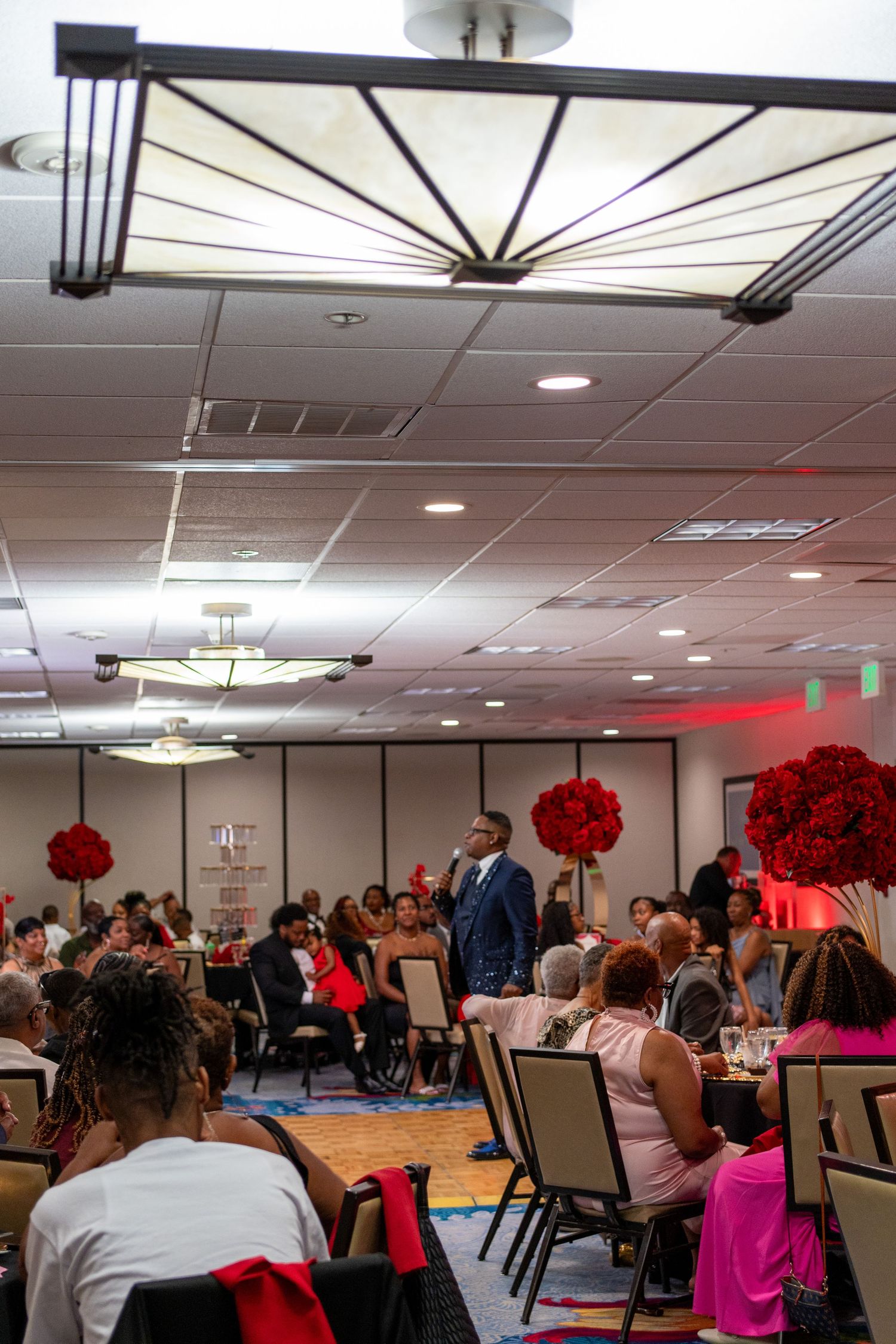 Elegant banquet hall event with guests seated at tables adorned with red floral centerpieces, while a man in a suit addresses the crowd.