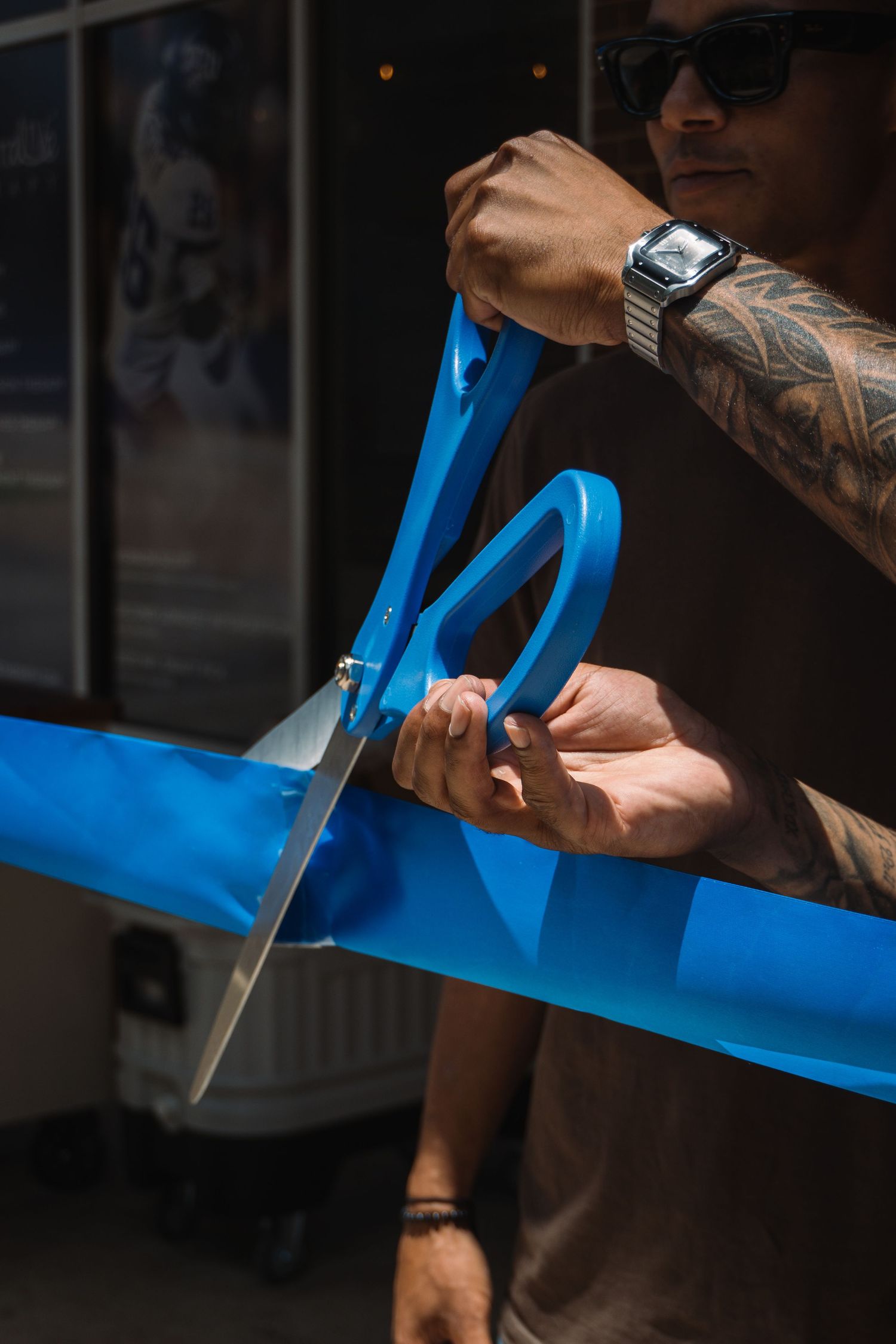 Tattooed man with smartwatch cutting blue ribbon with large scissors at a grand opening ceremony.