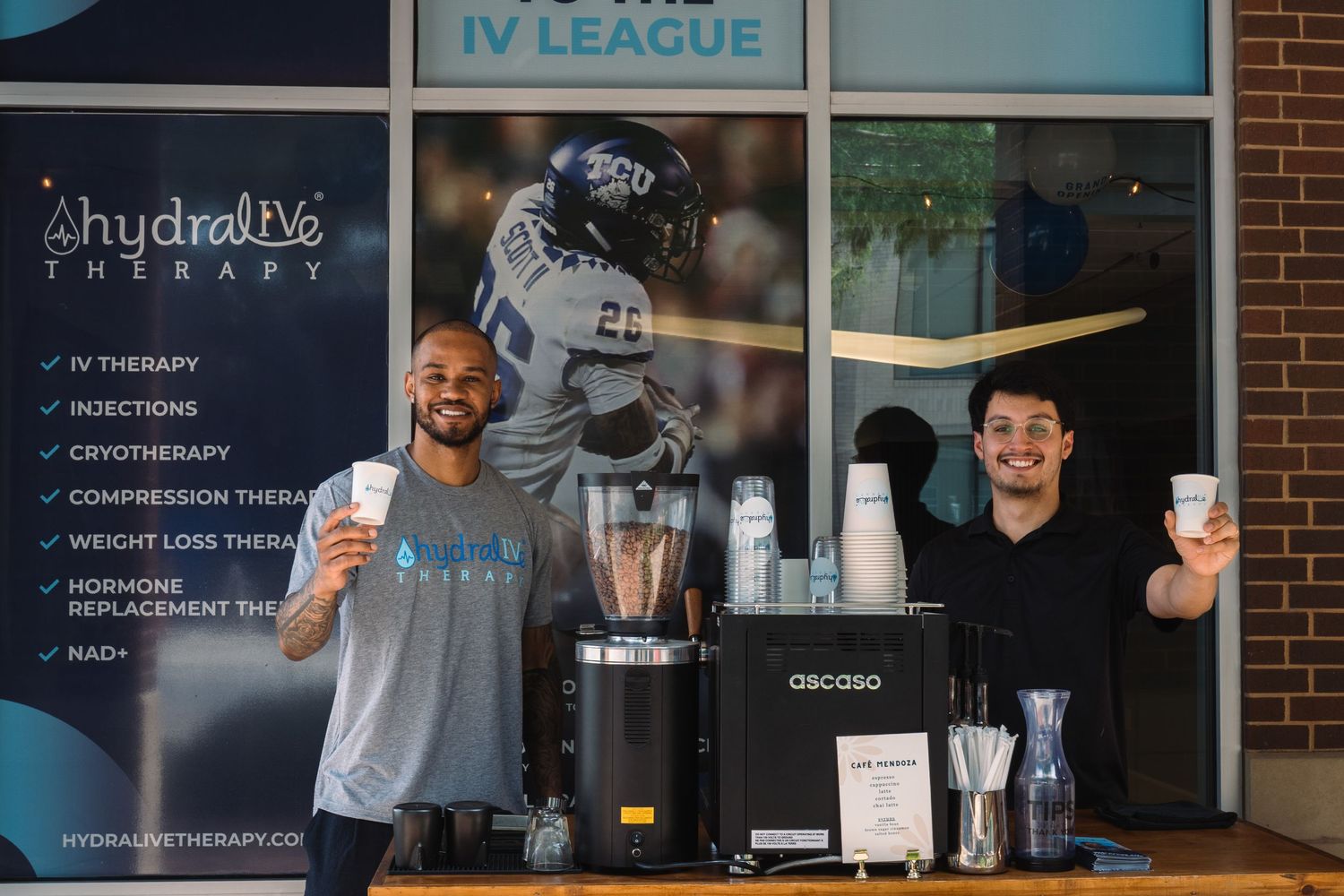 Two men smile behind a coffee and blender setup at HydraLyve Therapy IV League wellness event.