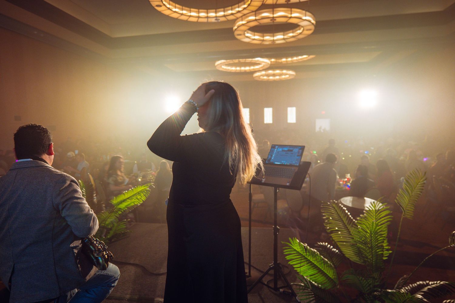 Woman in black dress speaks on stage at an elegant event with circular chandeliers and an engaged audience.