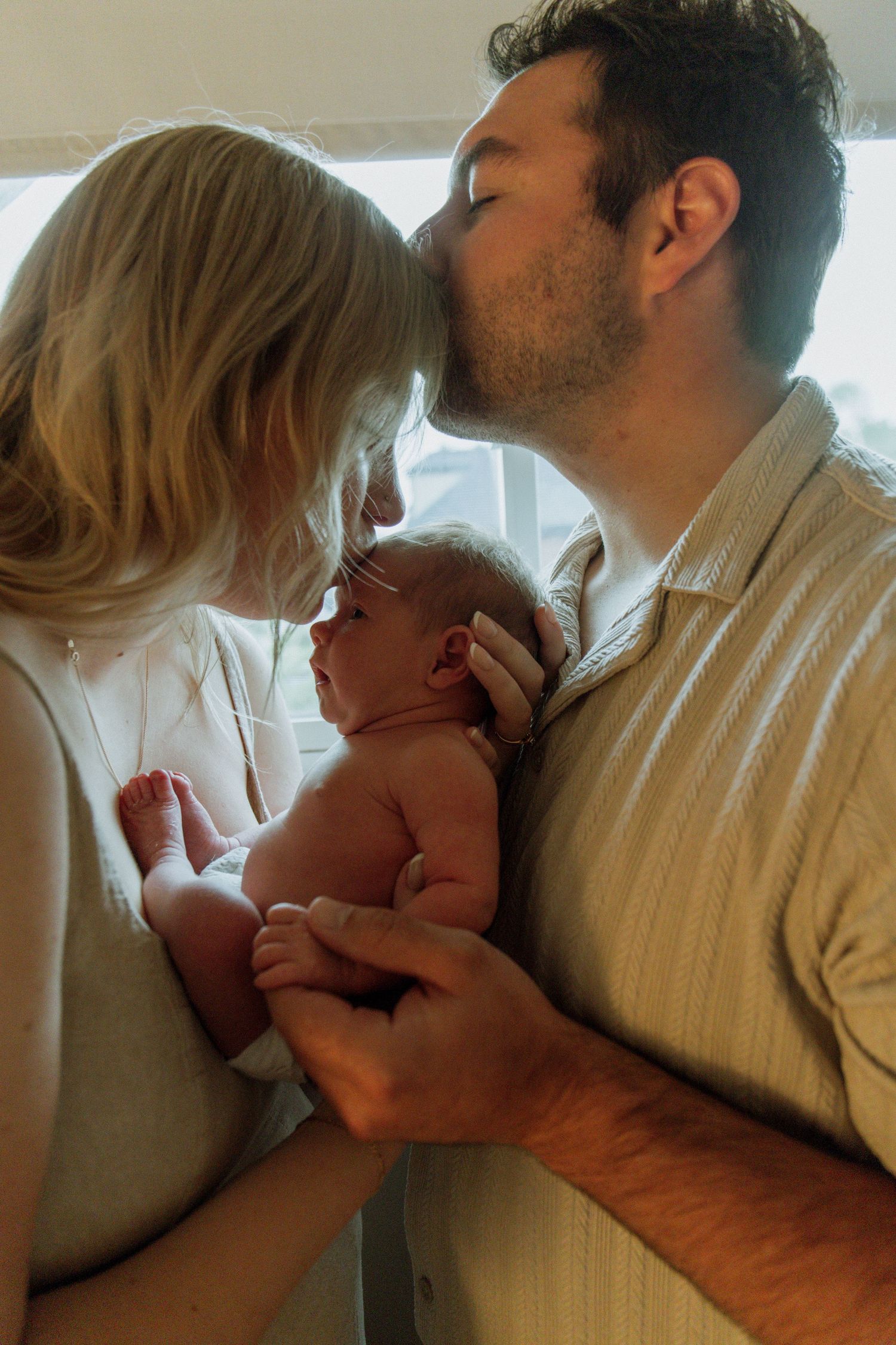 Kawartha Lakes Newborn Photography Session.
Mom and dad holding newborn baby girl between them. Dad kissing moms forehead. Mom kissing baby