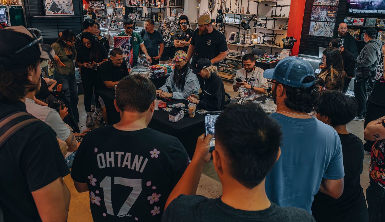 Crowd gathers around seated group at indoor trading card event.