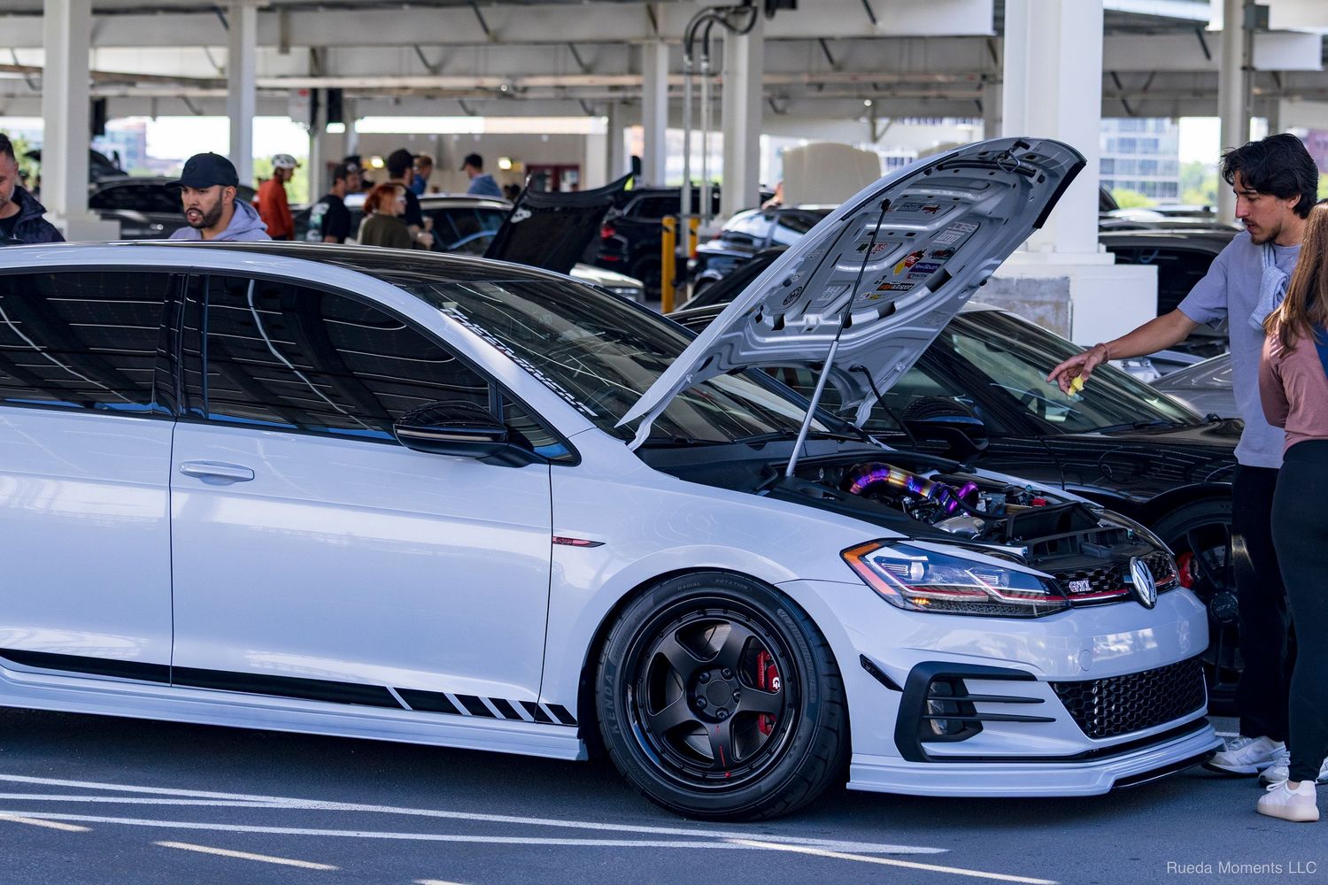 A modified white Volkswagen GTI with open hood and purple engine bay at a car show.