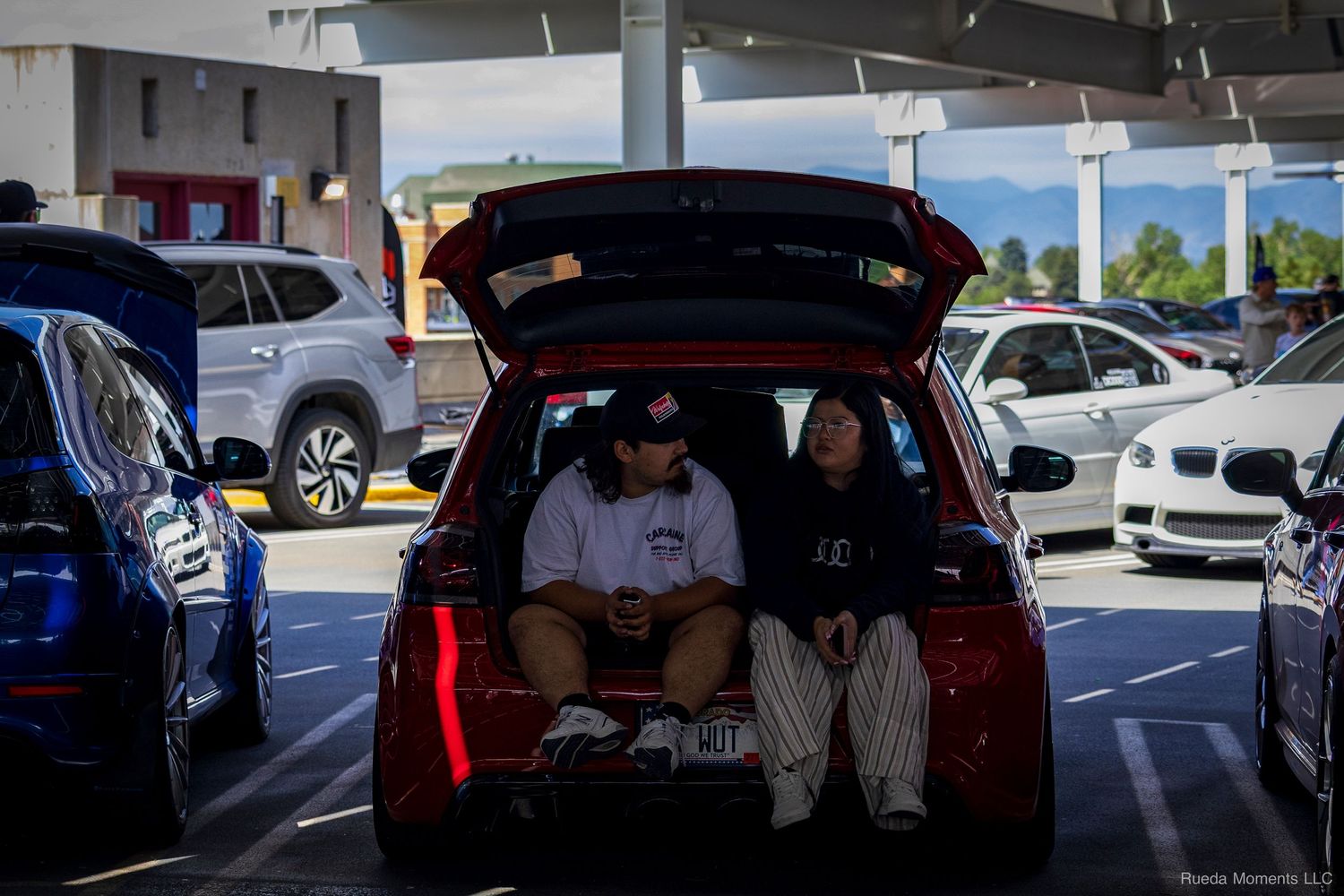 Two young people sitting in the open trunk of a red hatchback at an outdoor car meet event.