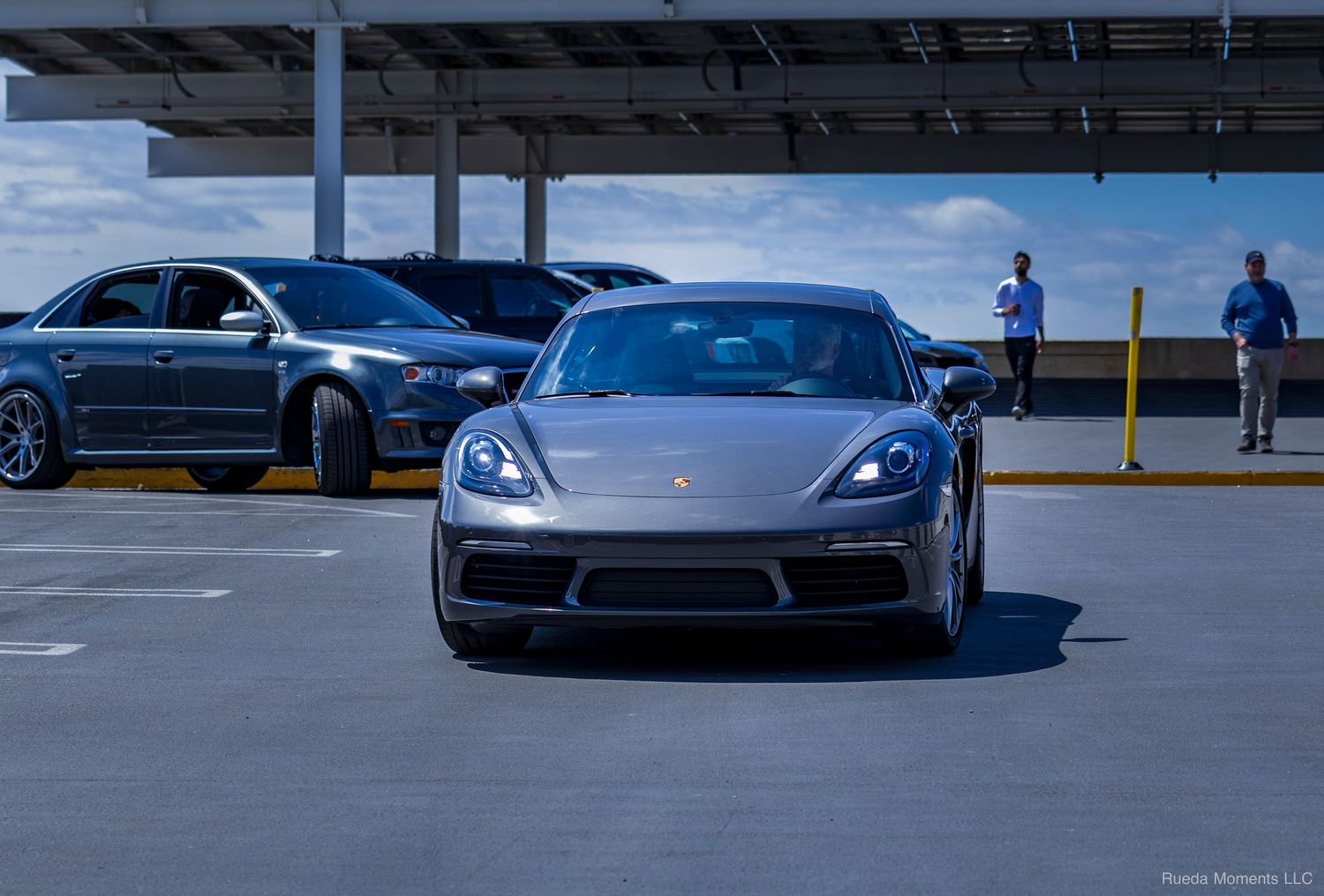 Silver Porsche Panamera driving through a sunny rooftop parking lot at a car meet.