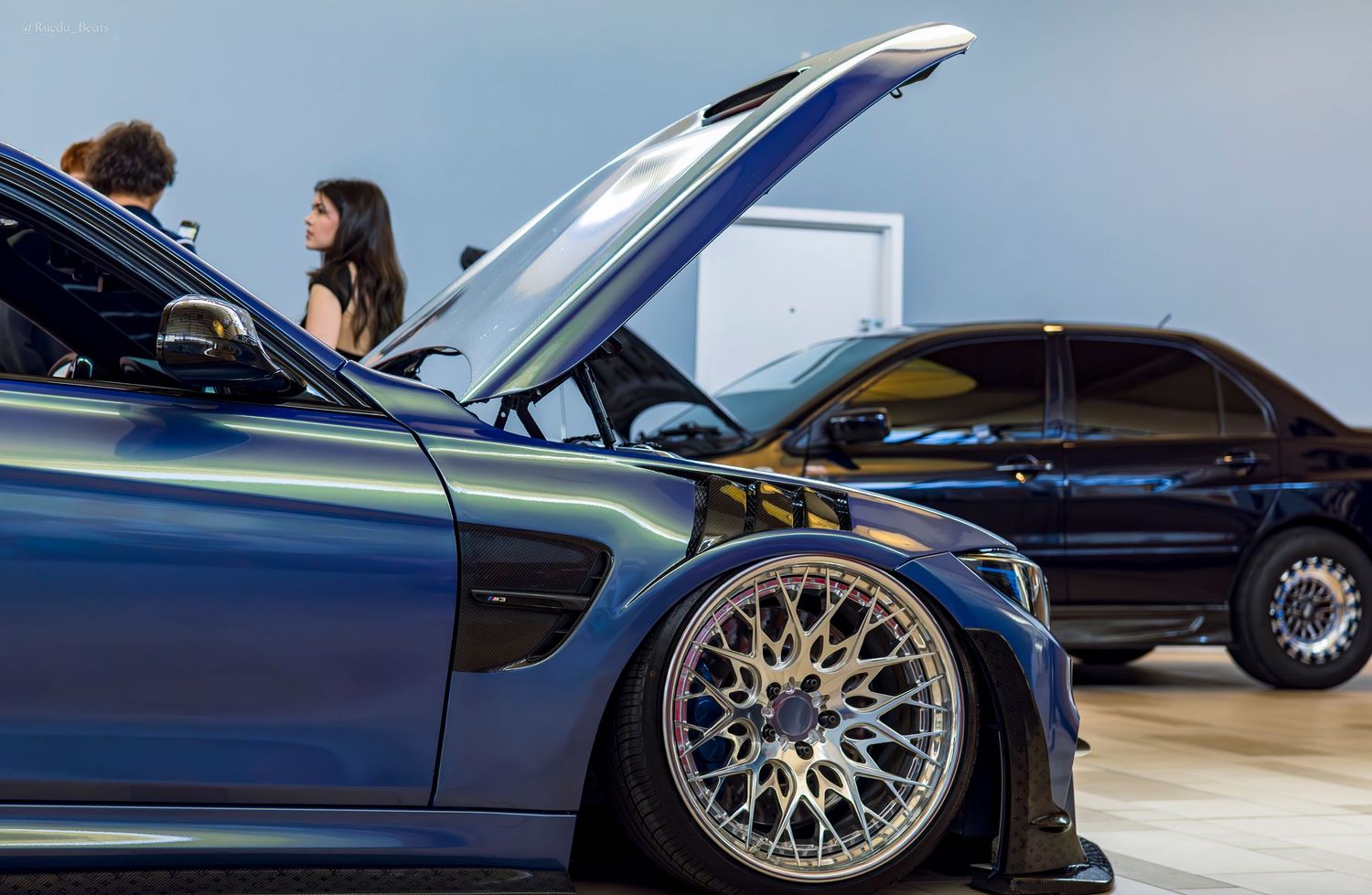 Two modified BMW cars with open hoods on display at an indoor automotive show.
