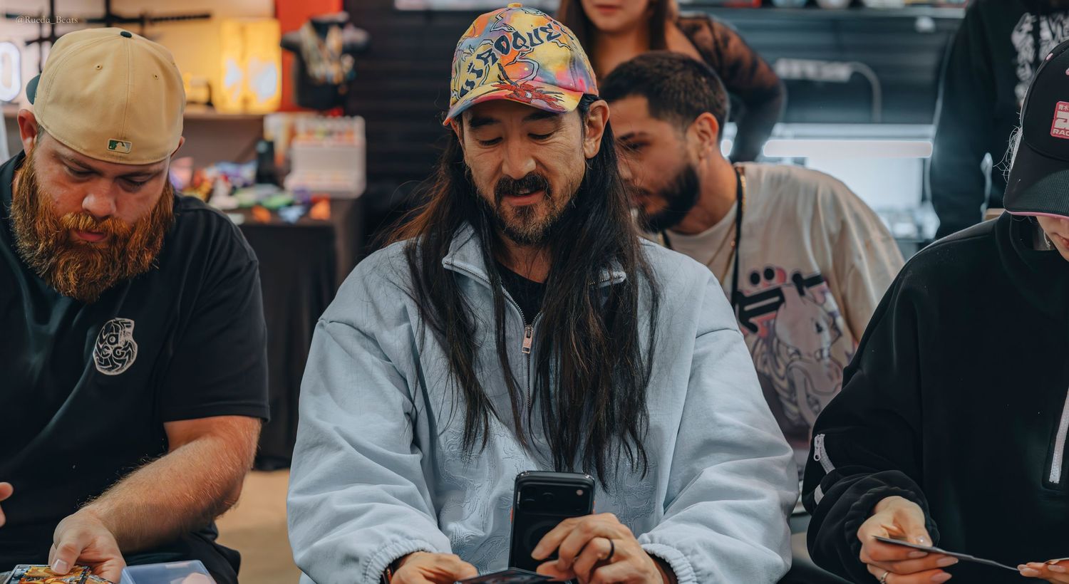 Long-haired man in colorful cap focused on his phone while opening trading cards at a store event.