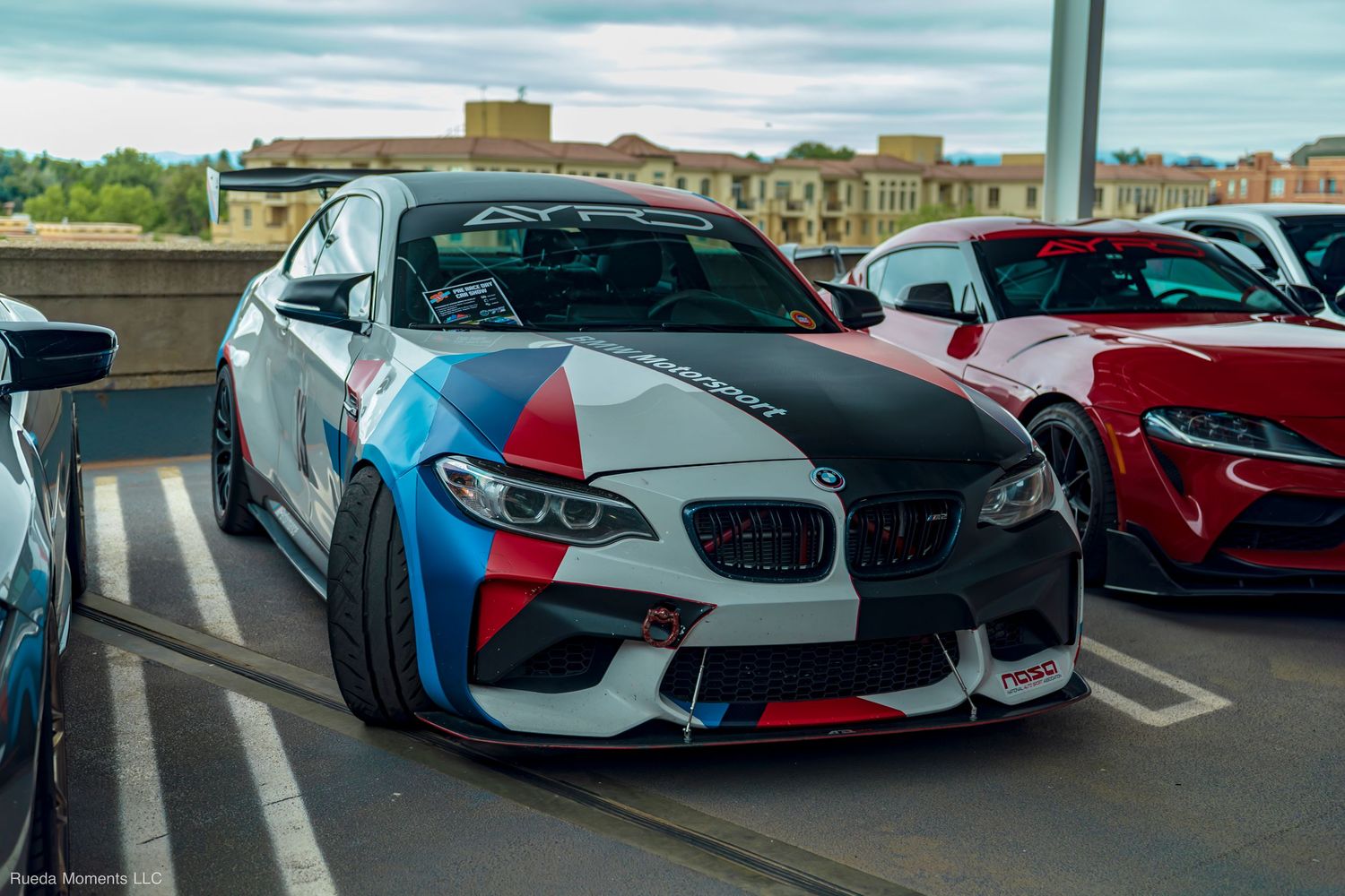 BMW M2 with aggressive widebody kit and M motorsport livery parked at an outdoor car show.
