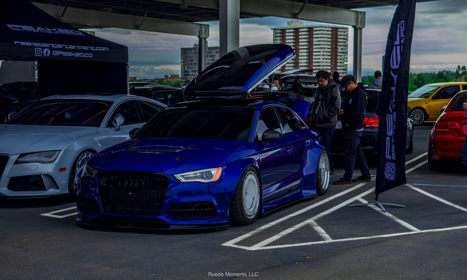 Modified blue Audi RS3 with roof box at outdoor car meet.