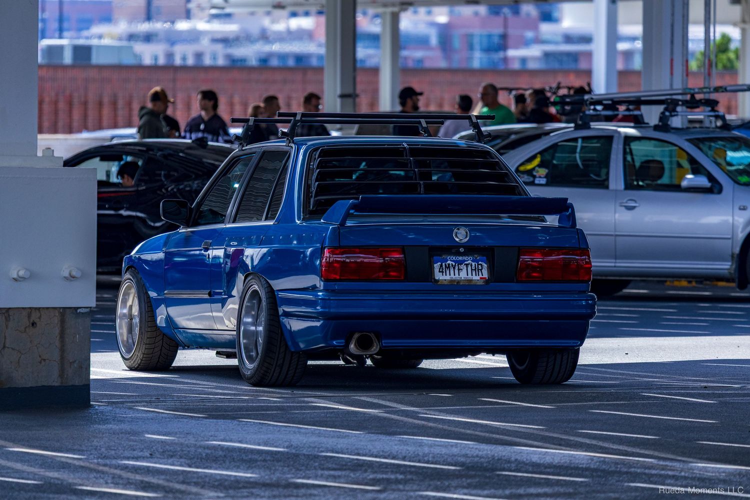 A customized blue BMW E30 with wide body kit and lowered stance at an outdoor car meet.