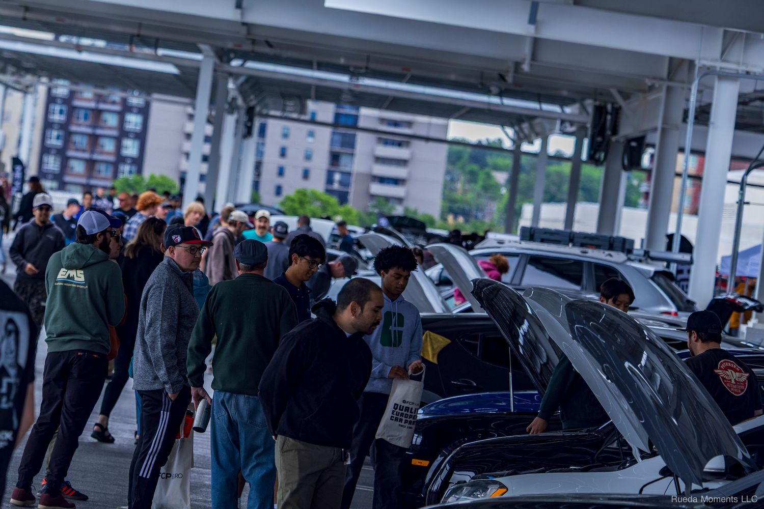 Crowd of car enthusiasts gathered around vehicles at an outdoor car show event.