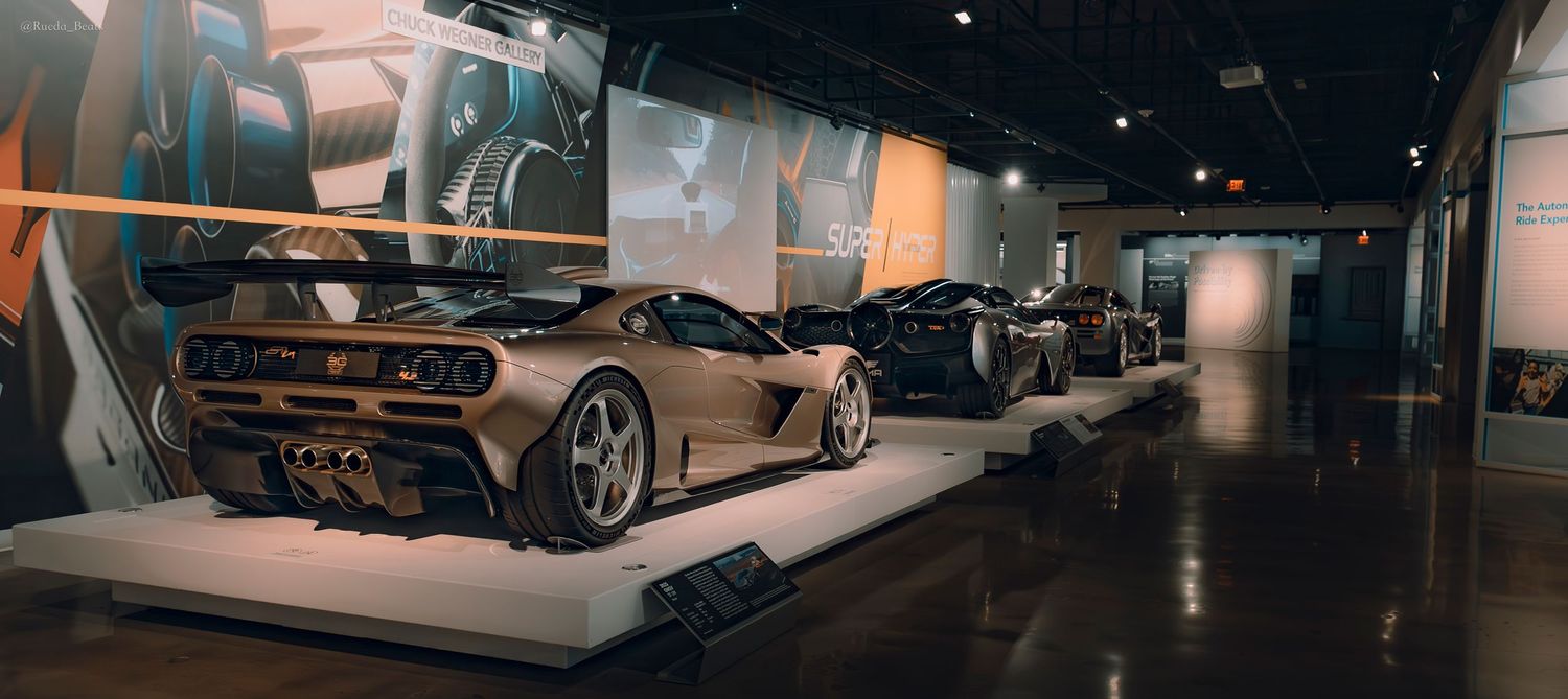 Wide angle view of exotic supercars on display in a dimly lit automotive museum gallery.
