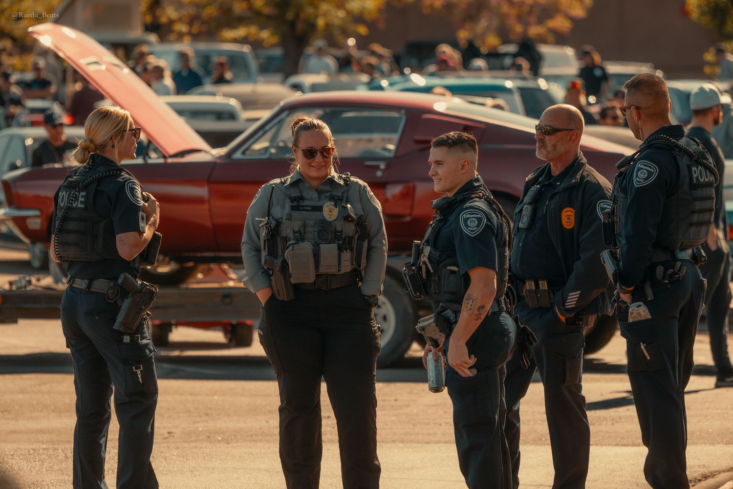 Group of police officers in uniform gathered in a parking lot near a red car at an outdoor event.